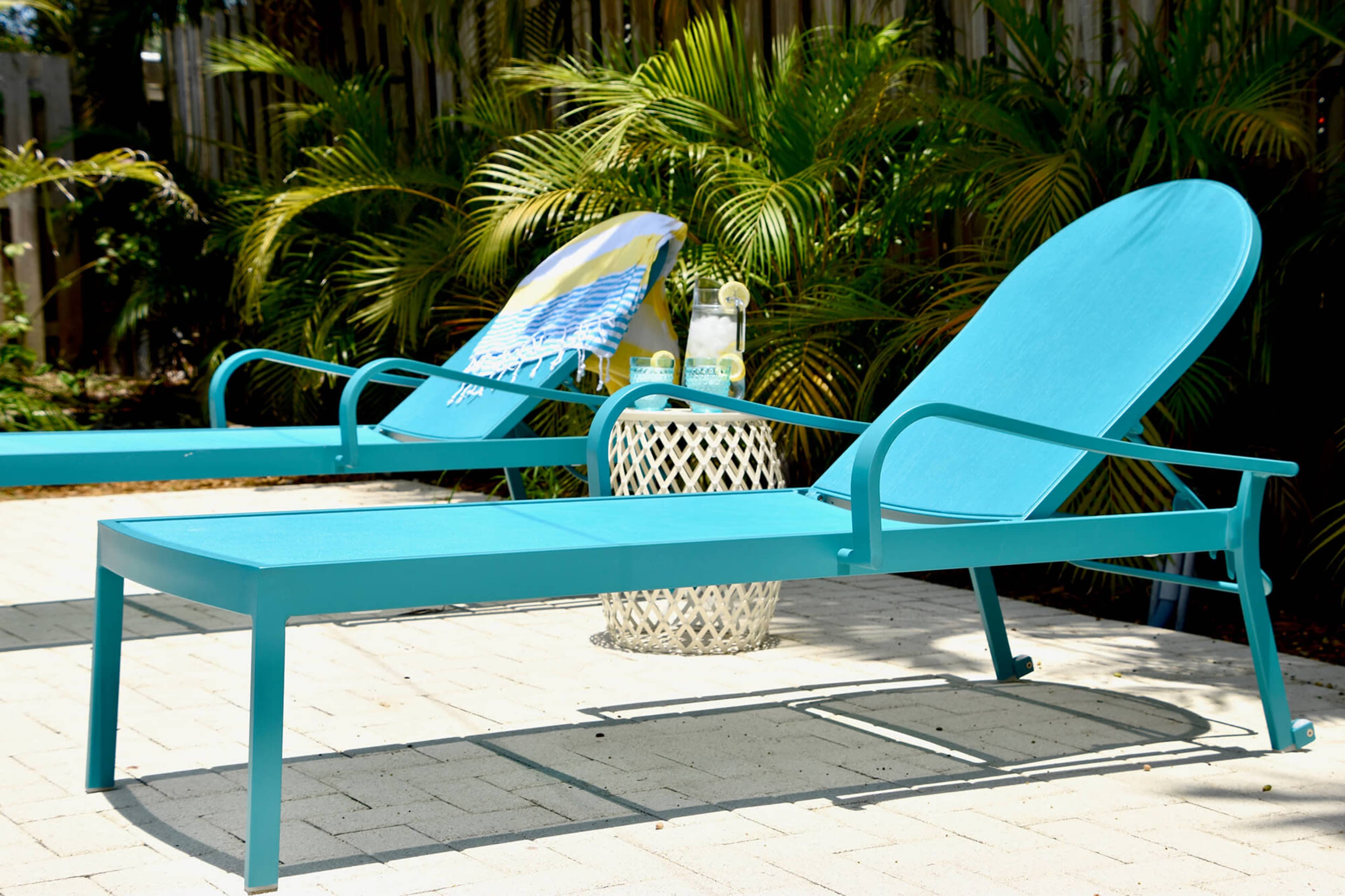Two turquoise lounge chairs are positioned on a patio surrounded by green plants, with a small patterned table holding two bottles of water nearby.