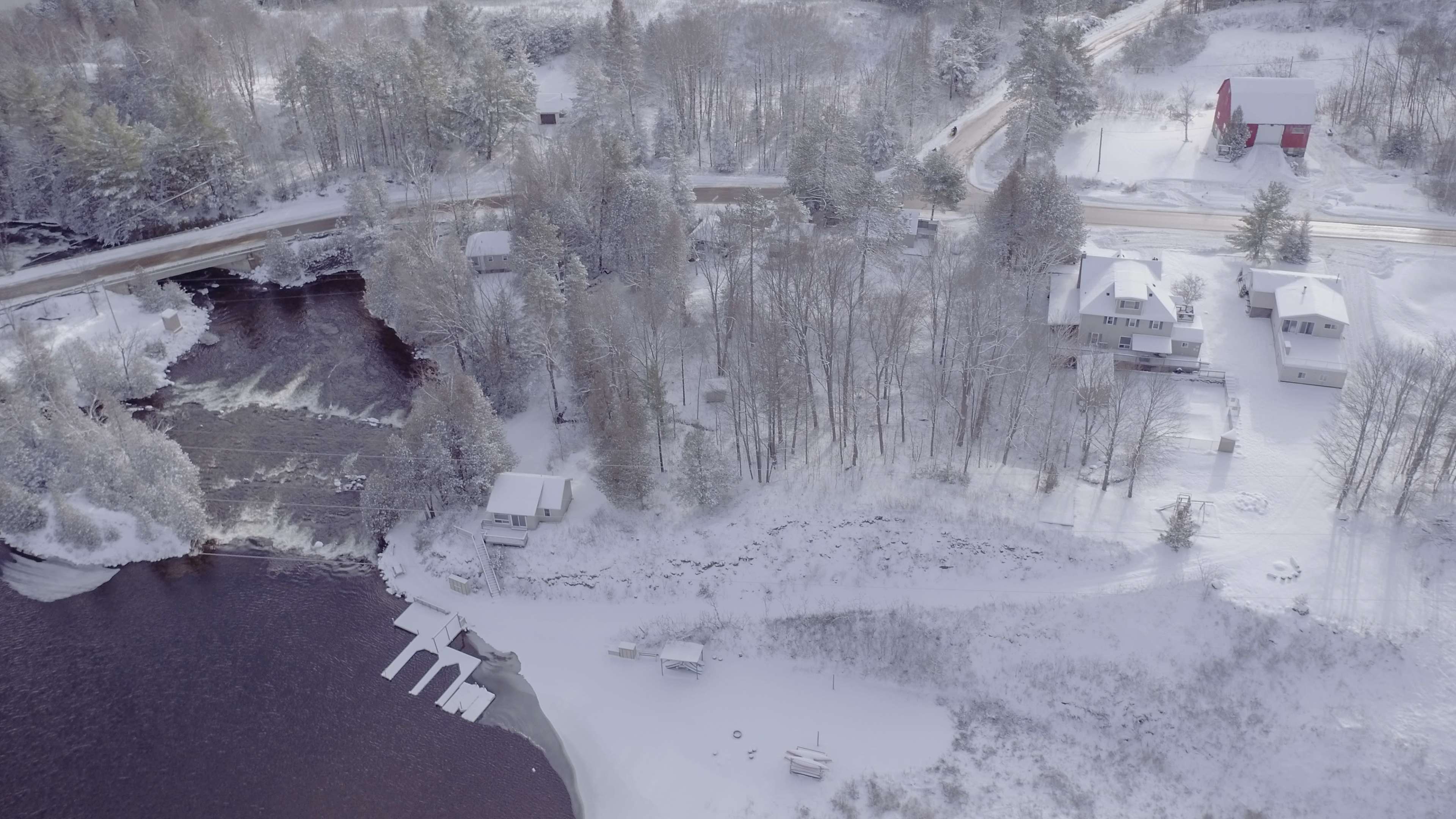 An aerial view of a snowy landscape featuring a river, several houses, and a road lined with trees.