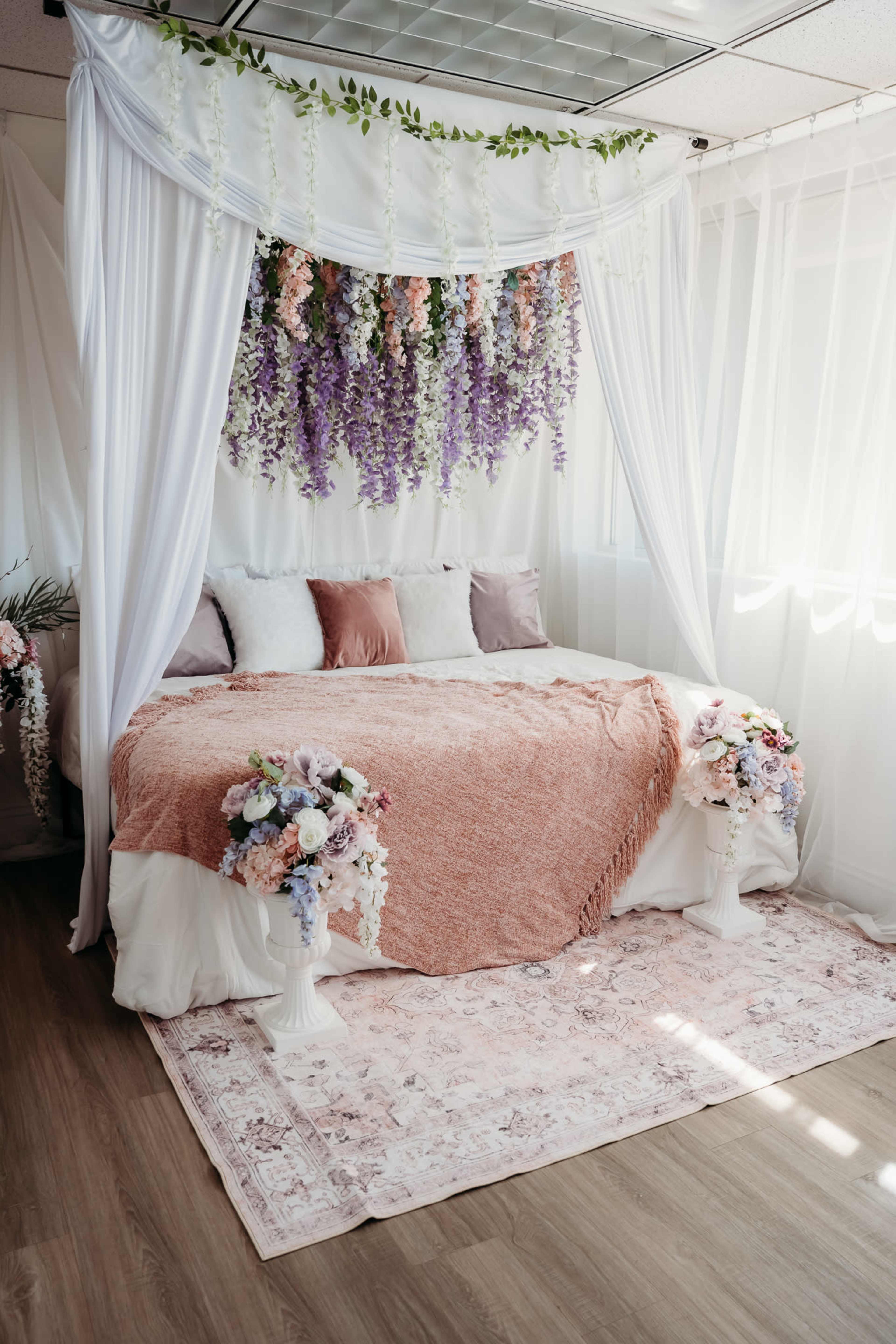 The image features a cozy bedroom with a bed draped in a white canopy, adorned with colorful floral arrangements and a soft rug beneath.