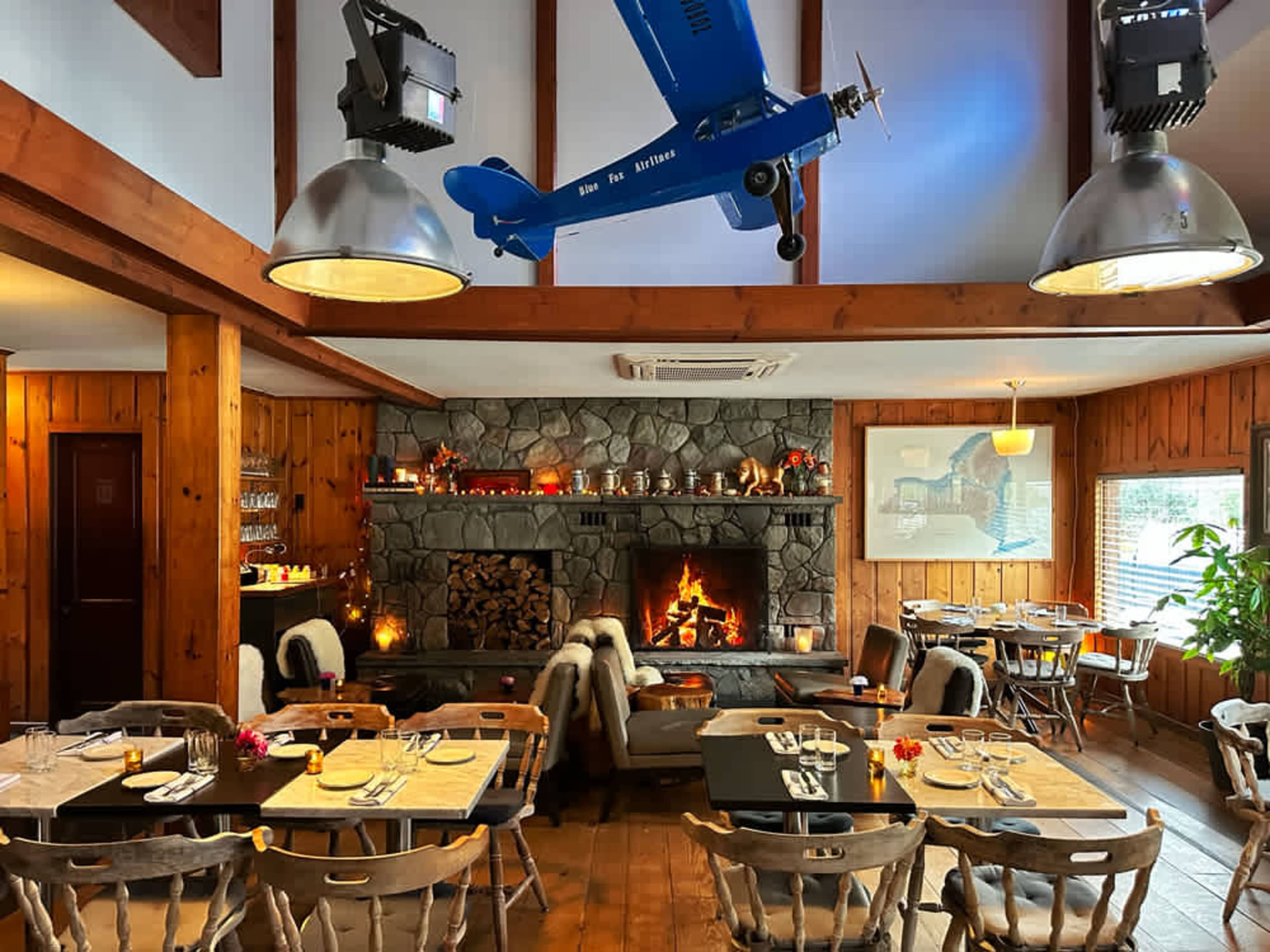 The interior of a restaurant features wooden walls, a stone fireplace with stacked logs, and tables set for dining under a model airplane hanging from the ceiling.