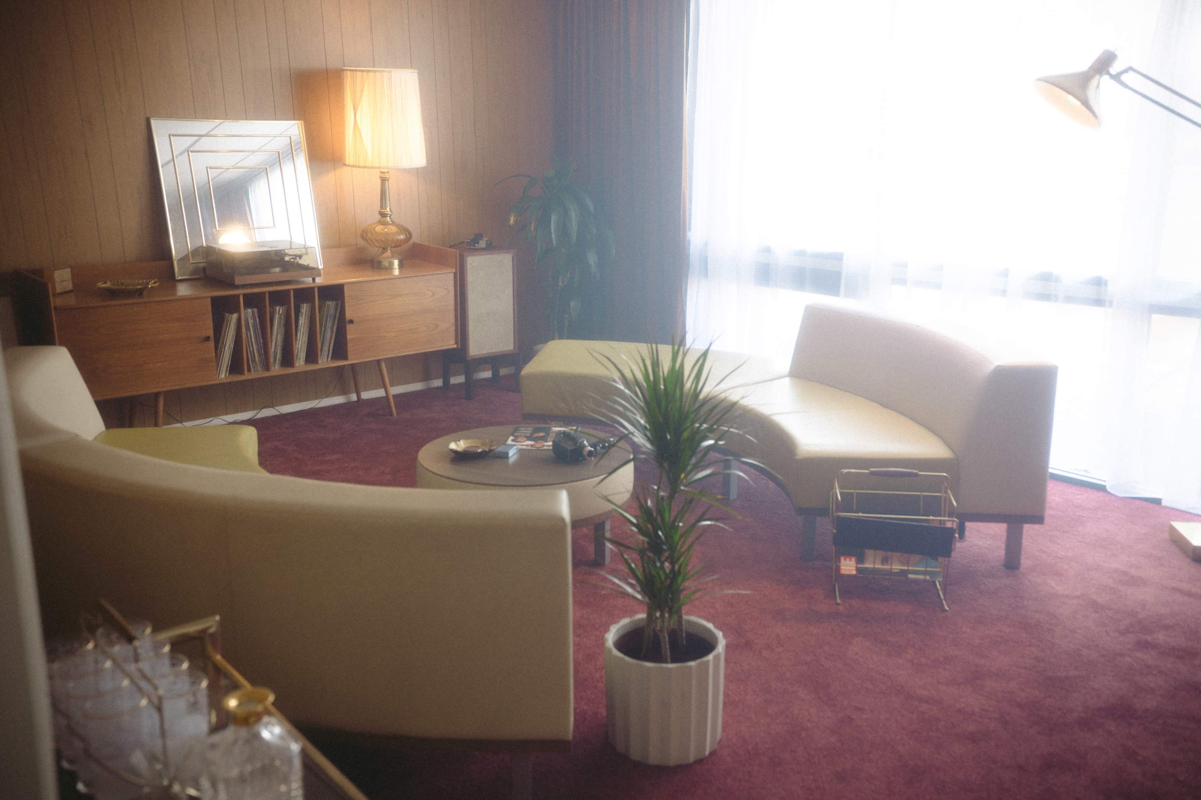 The image shows a mid-century modern living room with curved sofas, a round coffee table, and a decorative lamp, all situated on a carpeted floor.