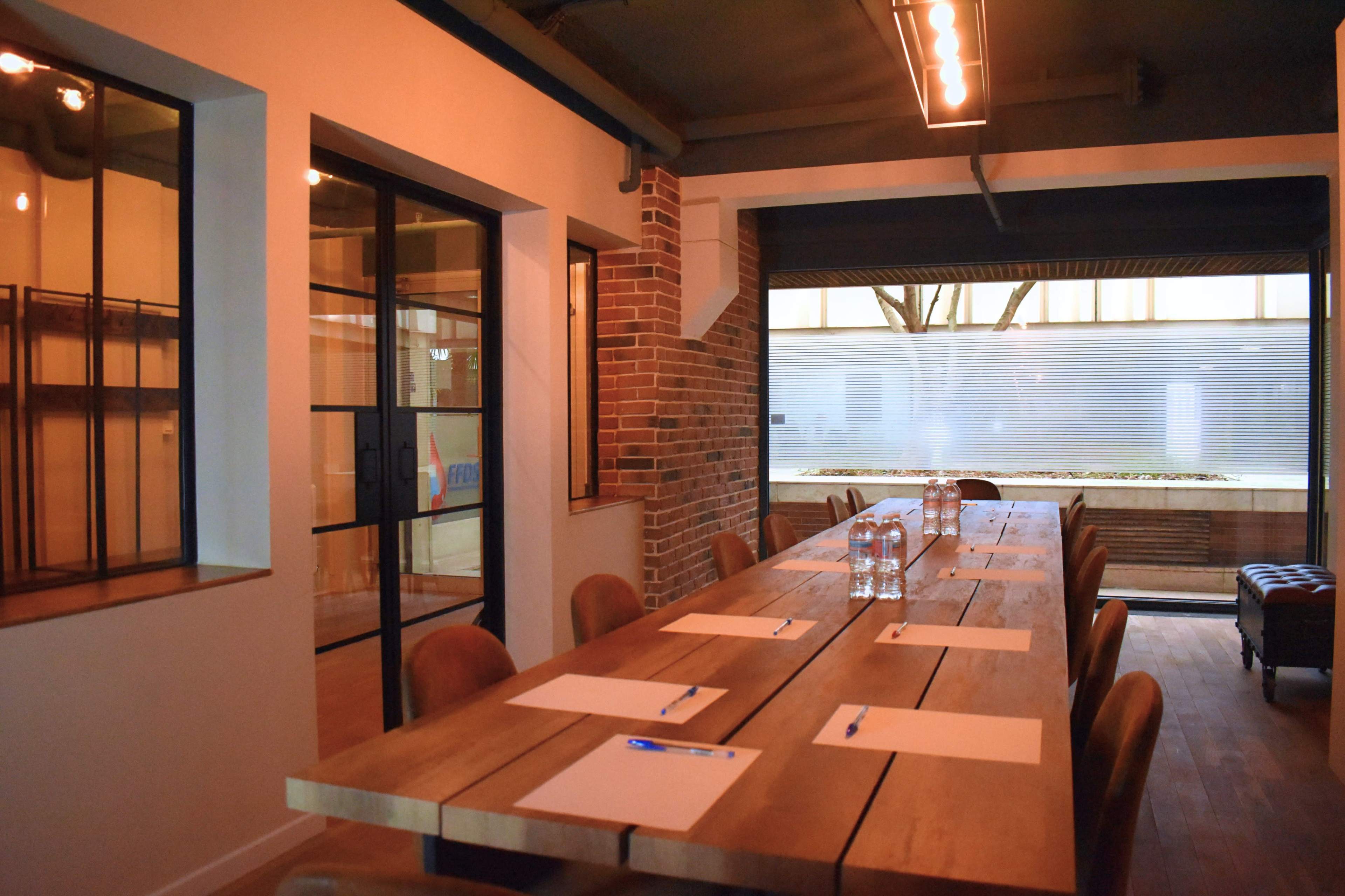 A large wooden conference table is set with paper and pens in a well-lit meeting room featuring brick and glass walls.