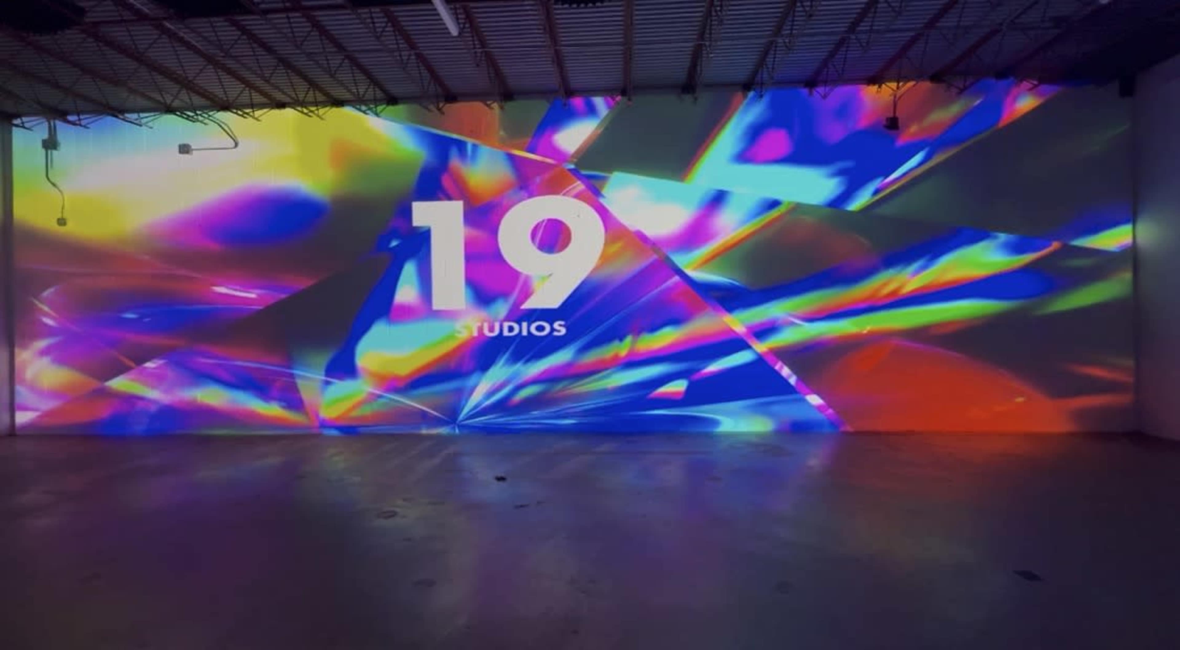 The image features a vibrant, multicolored projection on a large wall, displaying the text "19 STUDIOS."