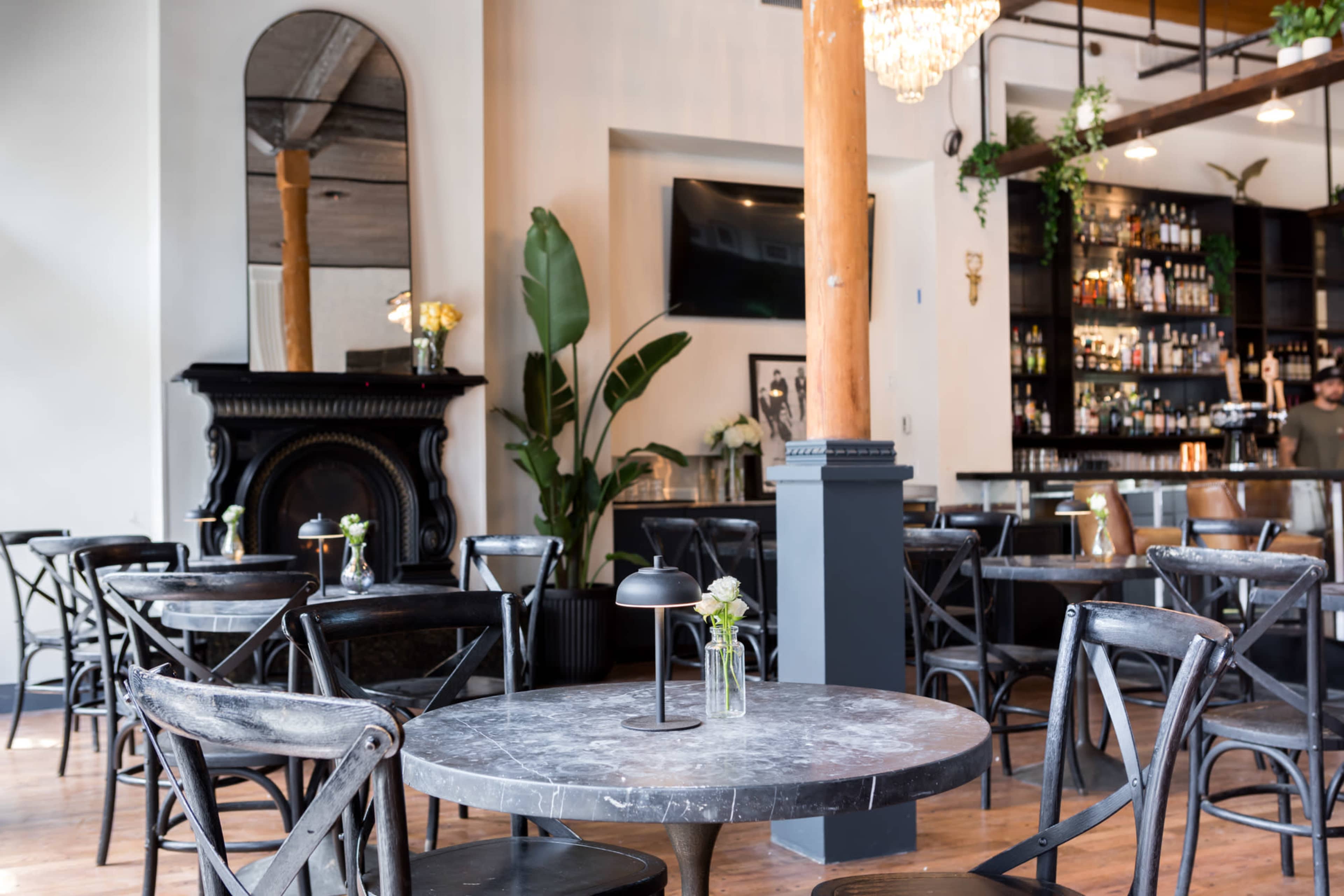 A modern café interior features wooden tables and chairs, elegant decor, a bar area, and a large mirror on the wall.