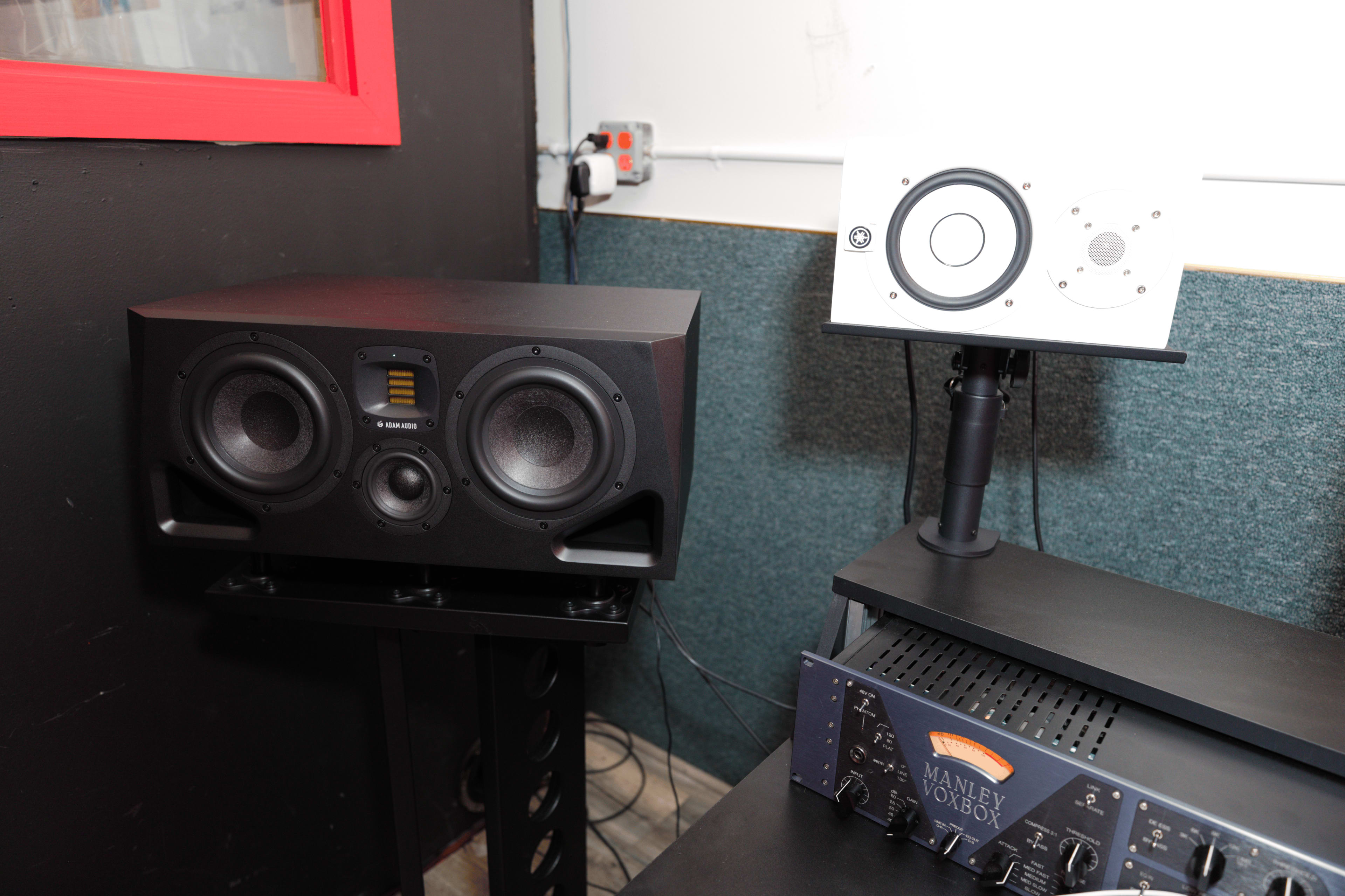 The image shows a pair of audio monitors on a stand next to a sound equipment rack with a visible audio processor.