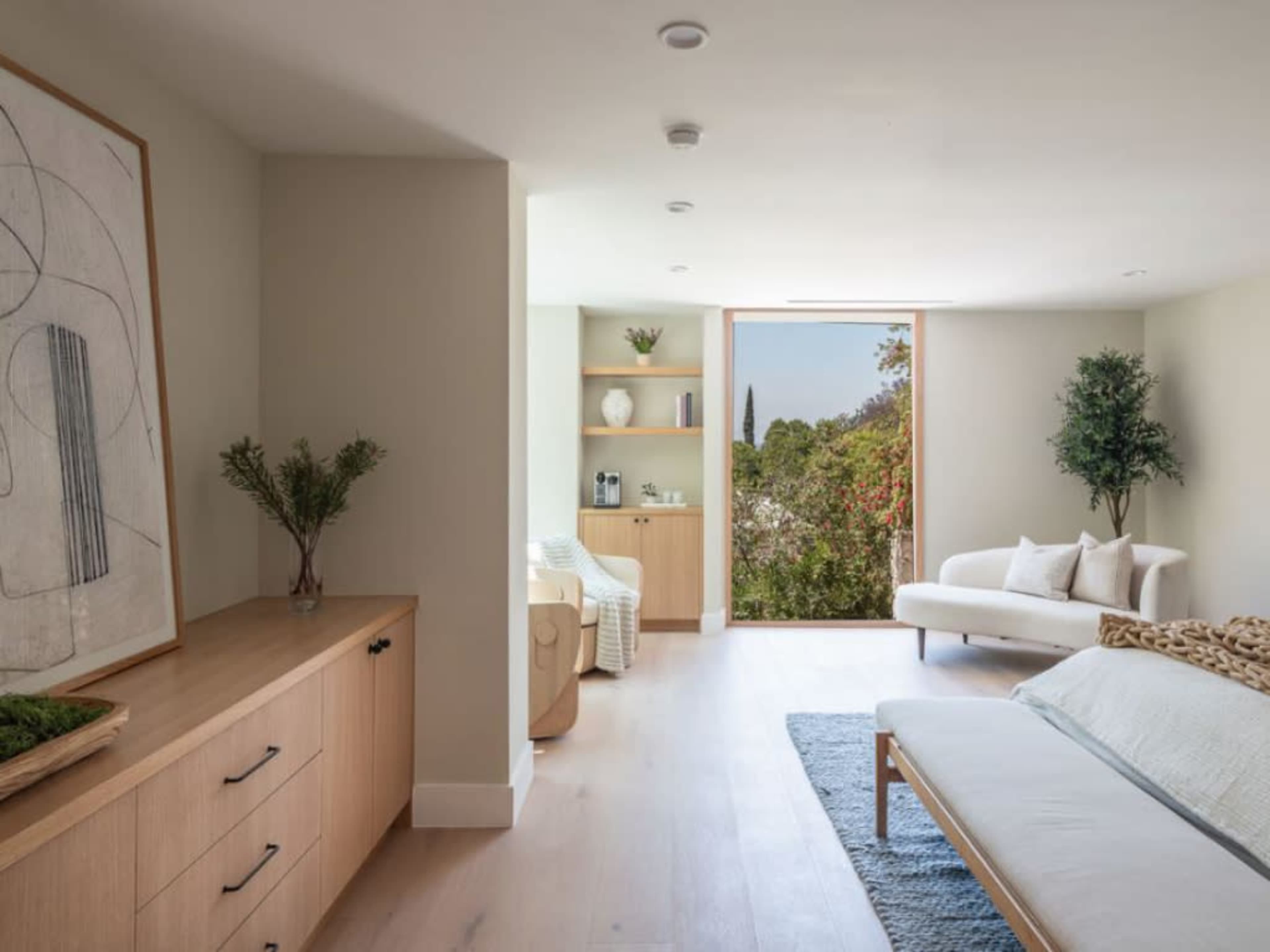 The image shows a bright, minimalist living room featuring a sofa, a bench, a wooden cabinet, and large windows with a view of greenery outside.