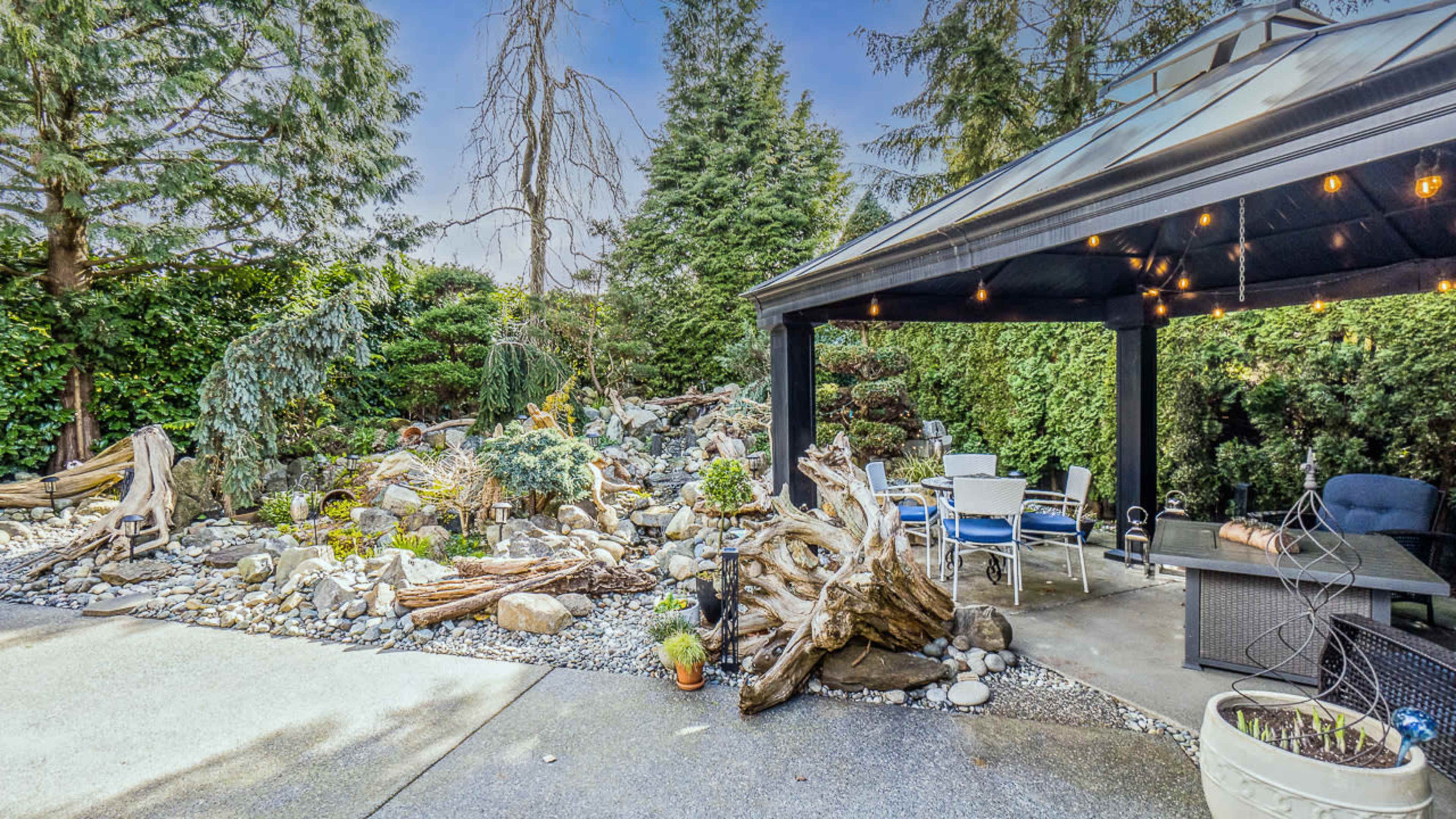 The image shows a landscaped garden featuring a rock and driftwood arrangement, with a gazebo on the right containing outdoor furniture.