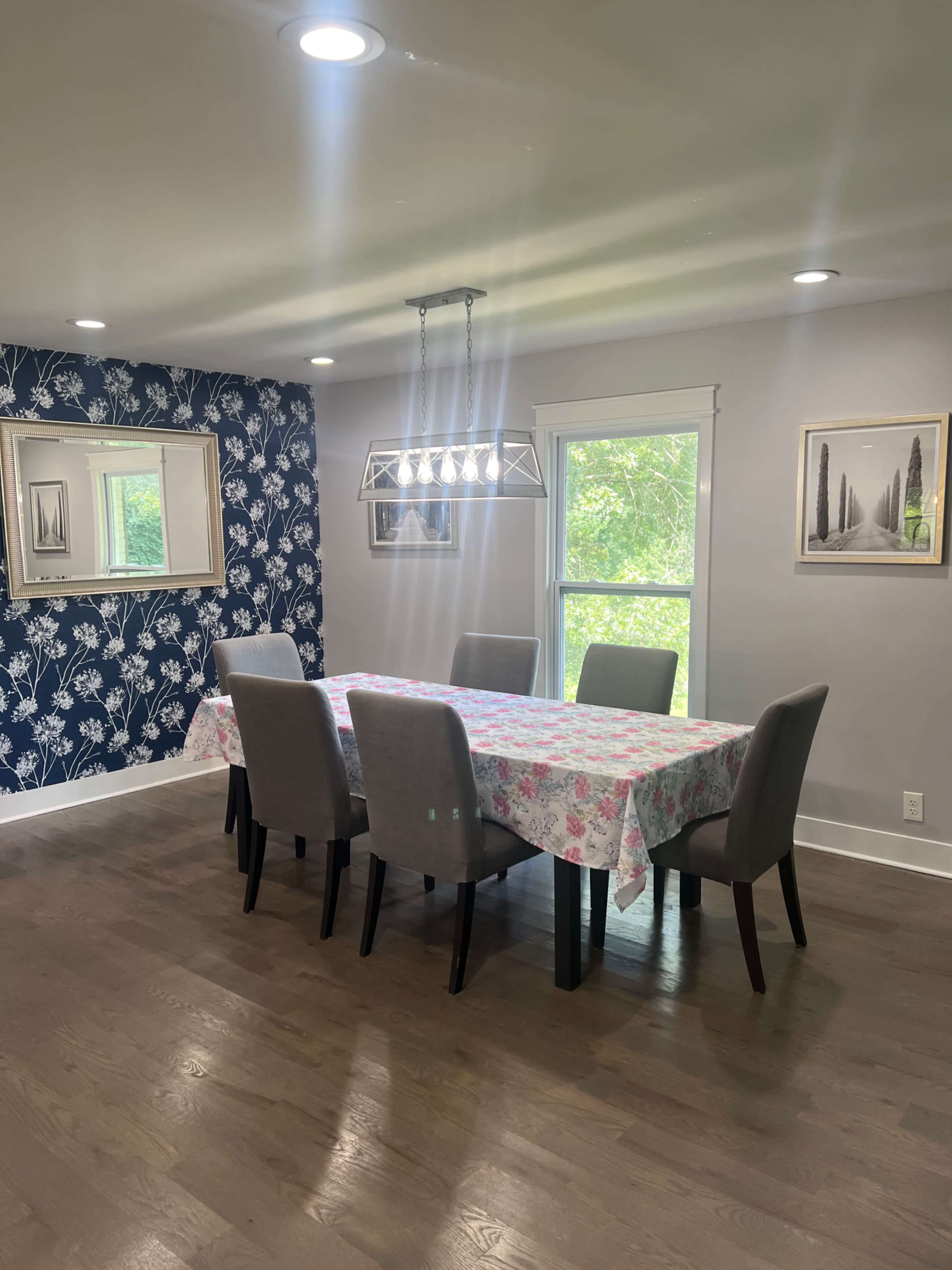 A dining room features a table covered with a floral tablecloth surrounded by six gray chairs, with a blue floral accent wall and a window overlooking greenery.