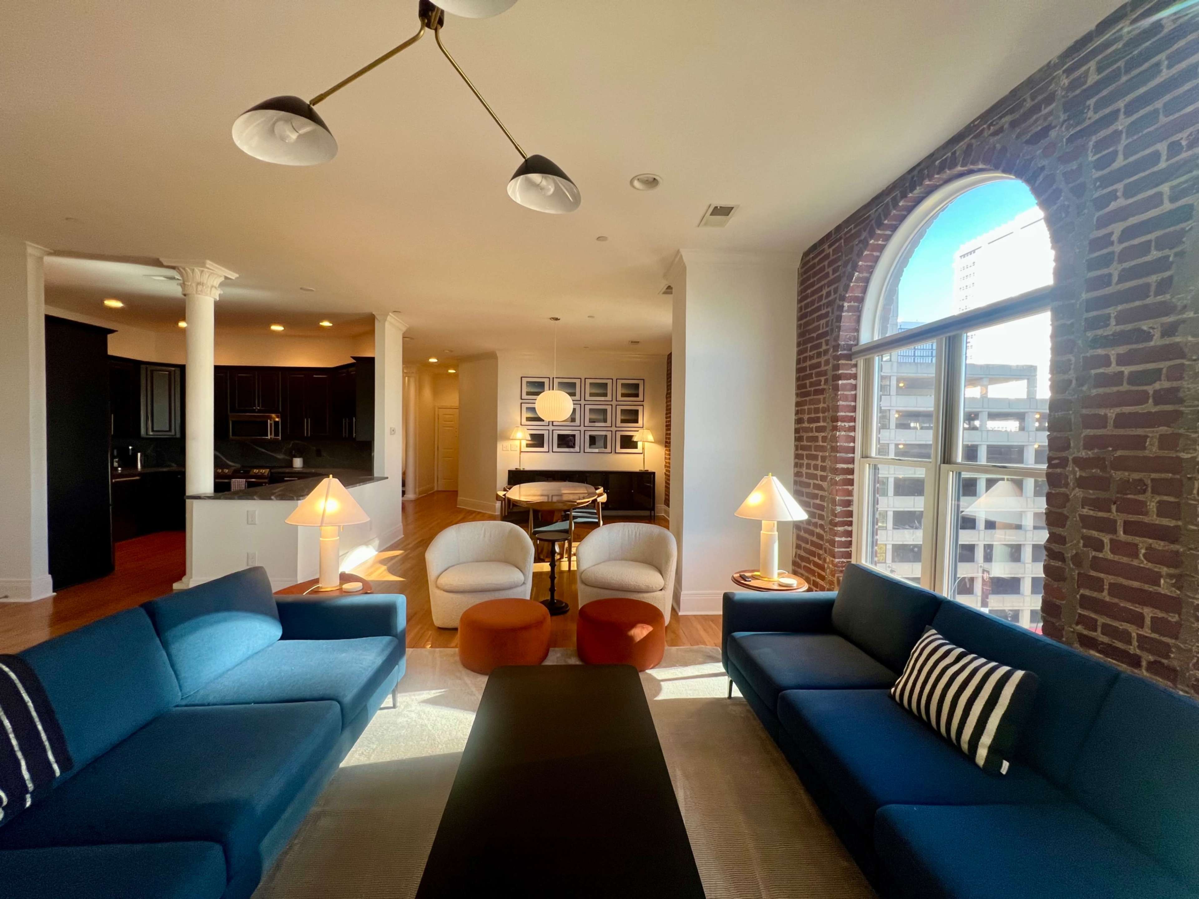 A spacious living room features two blue sofas, a black coffee table, and two accent chairs, with large windows showcasing a city view and exposed brick walls.
