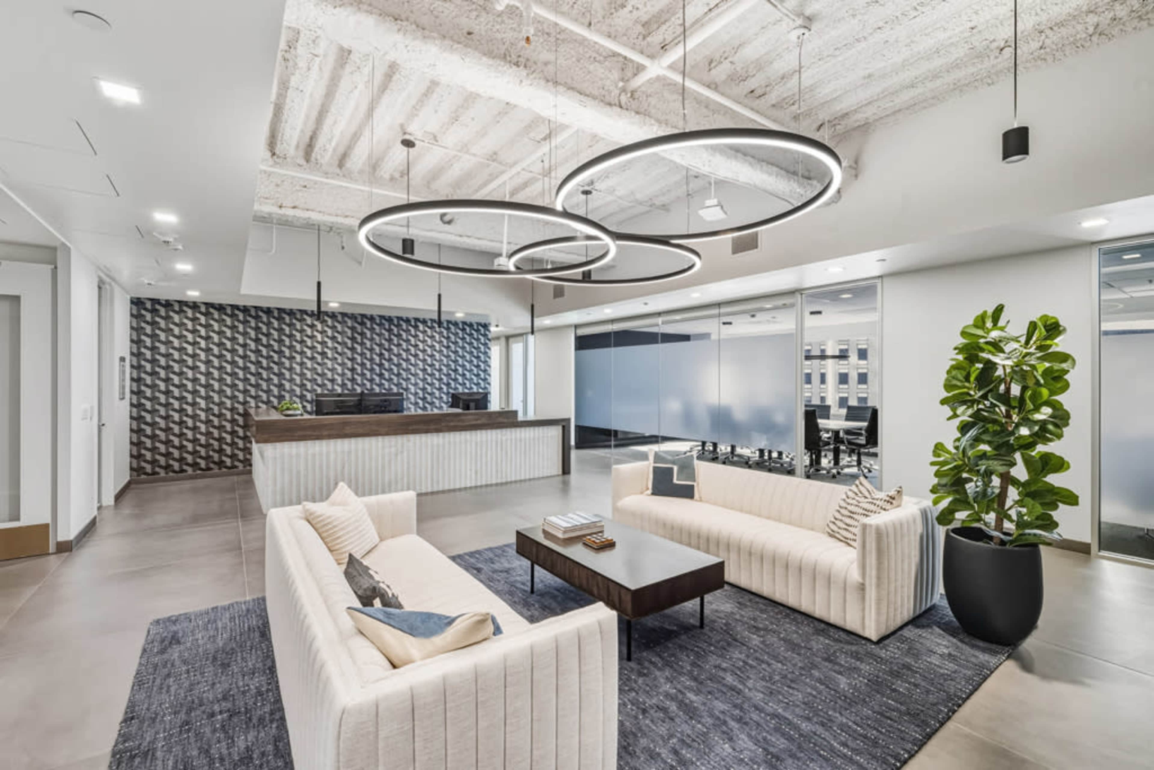 The image shows a modern office lobby featuring two white sofas arranged around a black coffee table, with a reception area in the background and a large decorative plant in a pot.