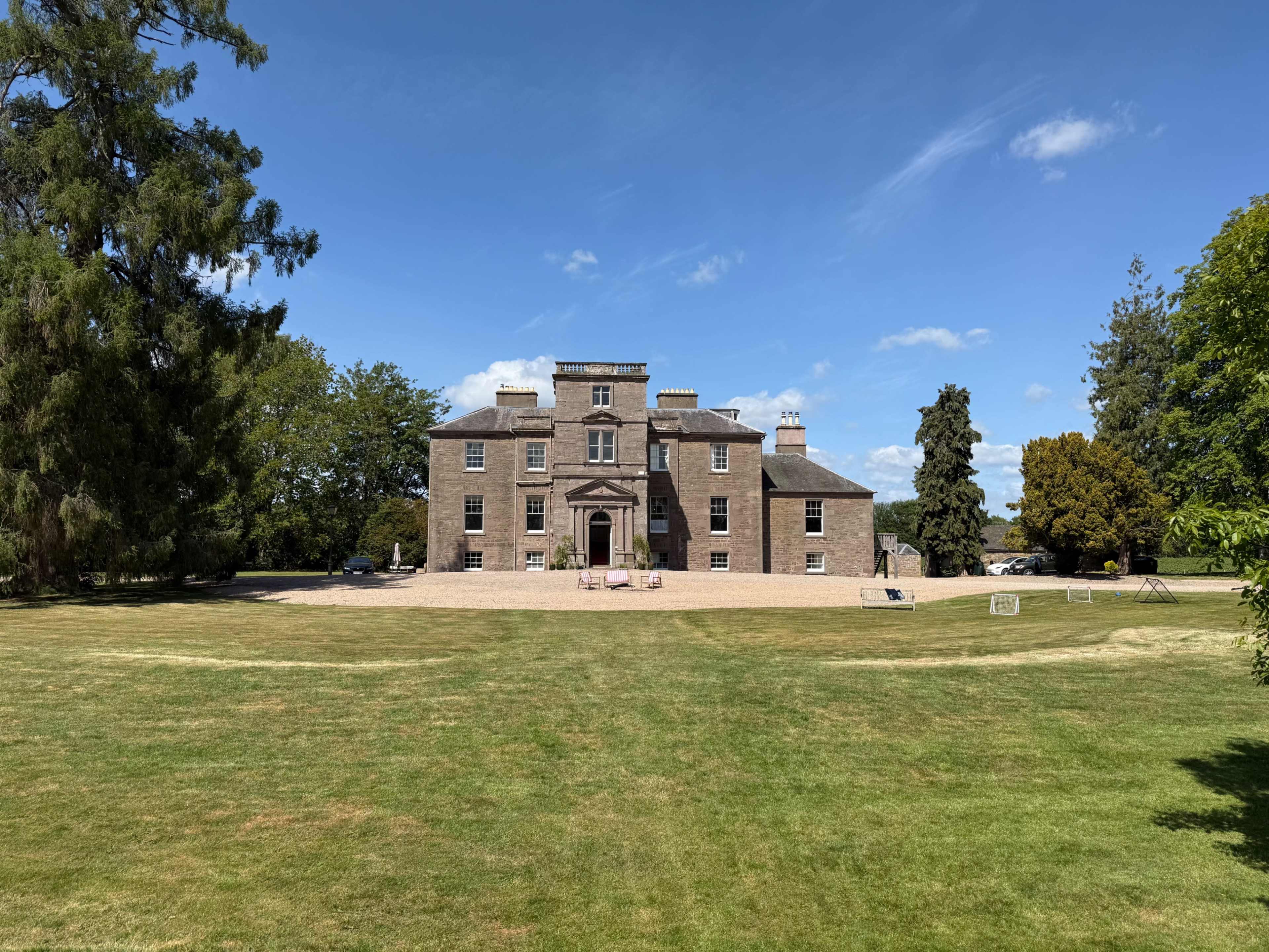 A large, historic brick mansion with a symmetrical façade and a spacious front lawn is set against a clear blue sky.