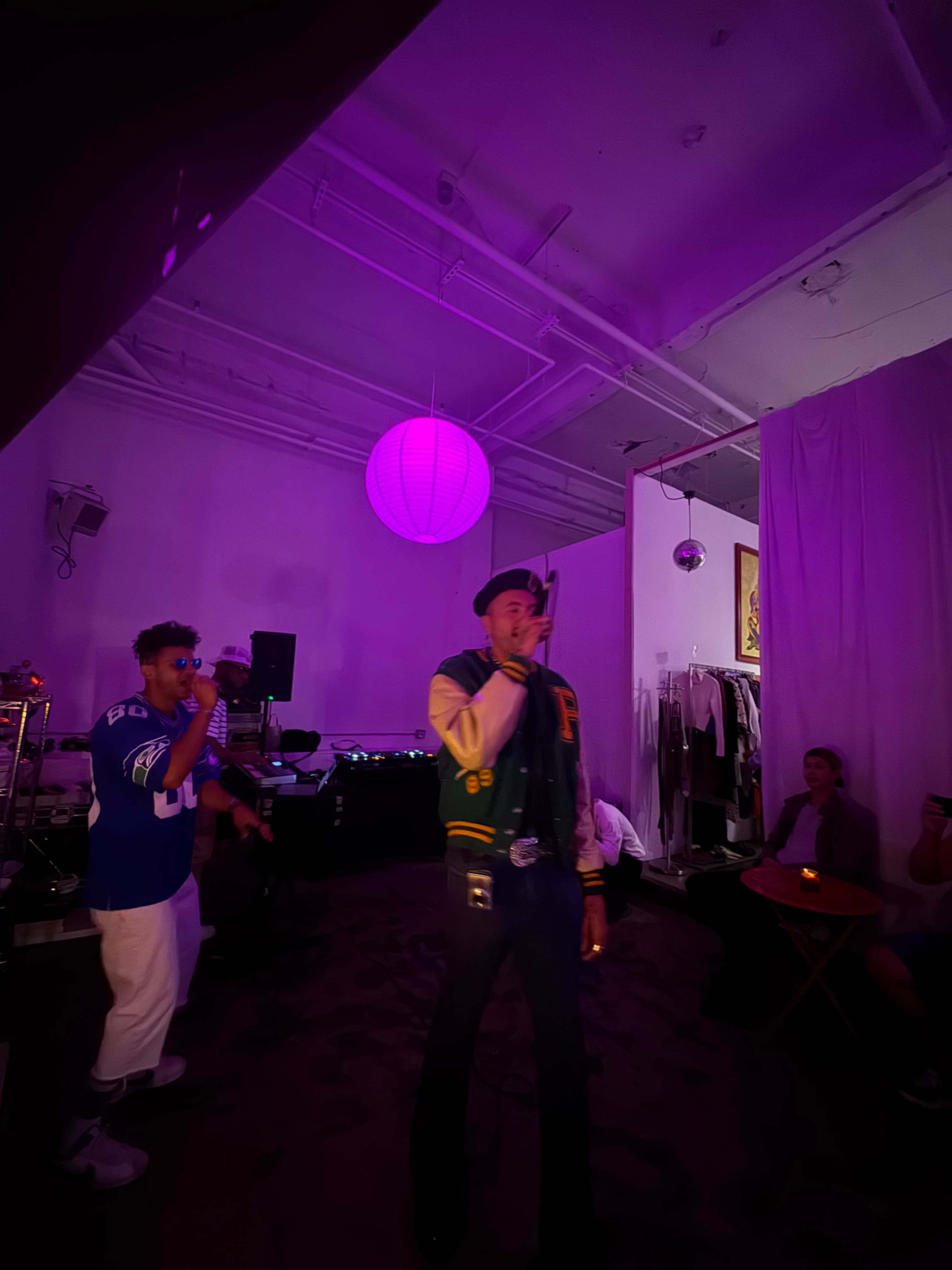 A performer raps on stage in a dimly lit room with purple lighting, while attendees sit nearby, enjoying the show.