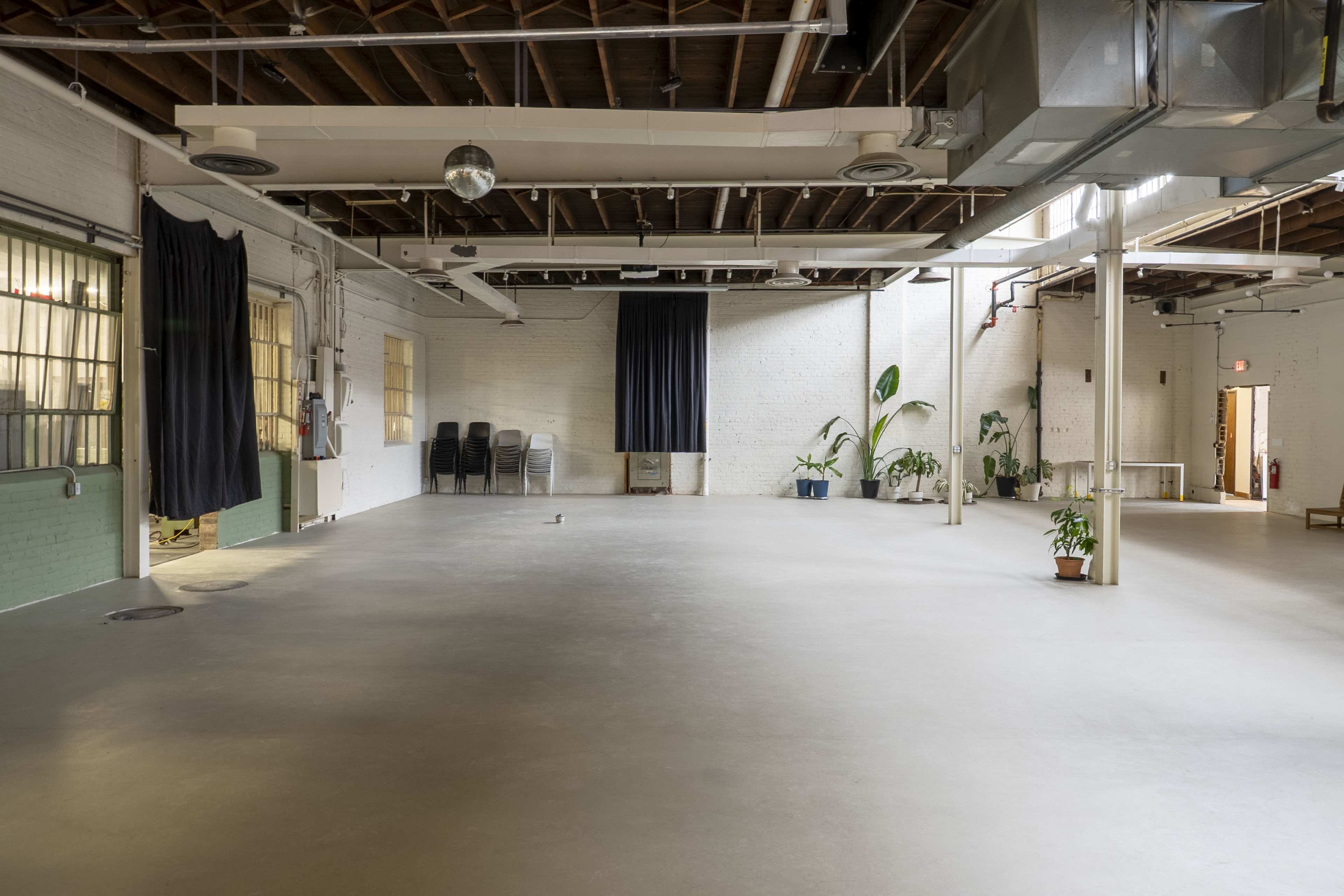 The image shows a large, open interior space with unfinished walls, large windows, and minimal furnishings, including a few potted plants and chairs stacked in a corner.