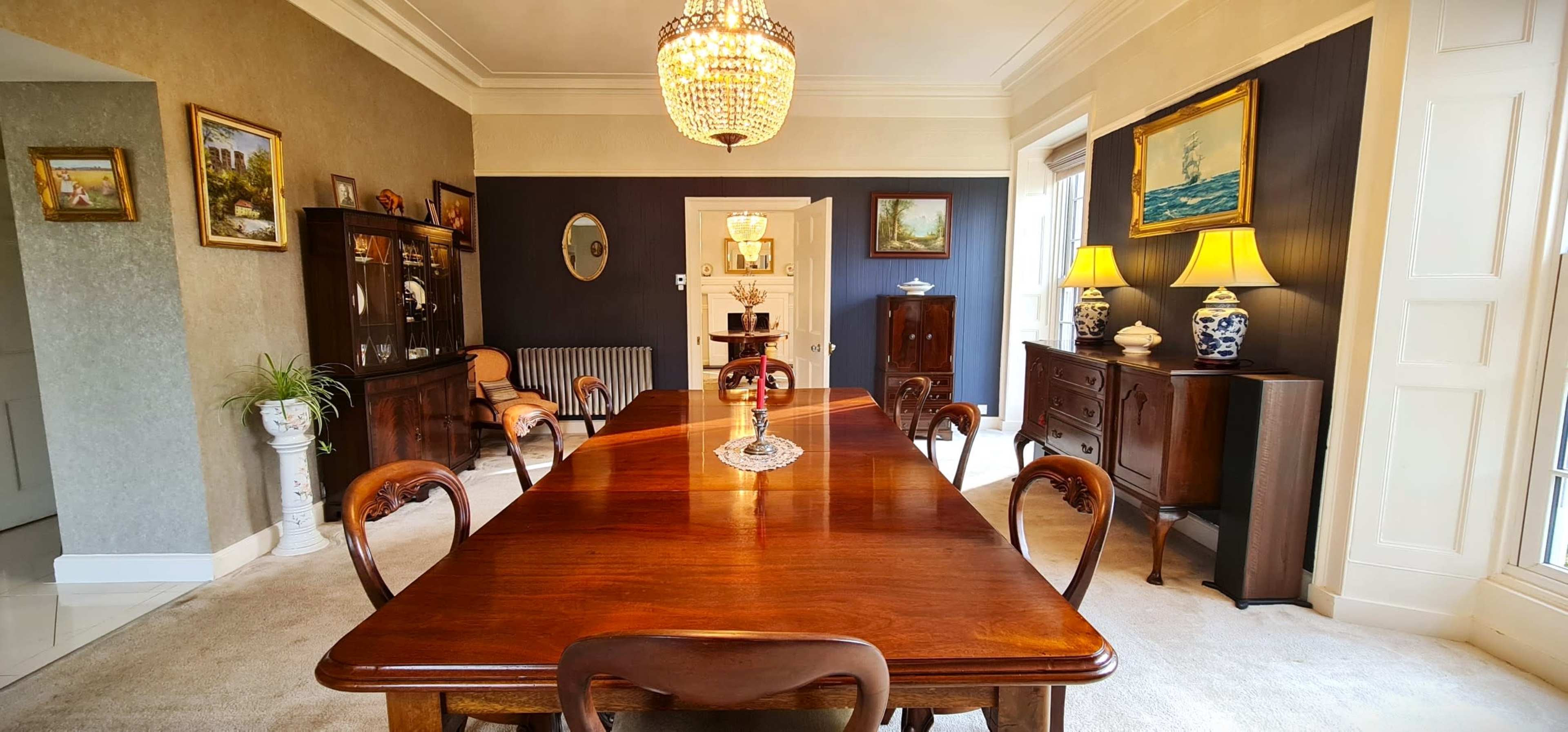 The dining room features a long wooden table surrounded by chairs, with decorative artwork on the walls and a chandelier overhead.