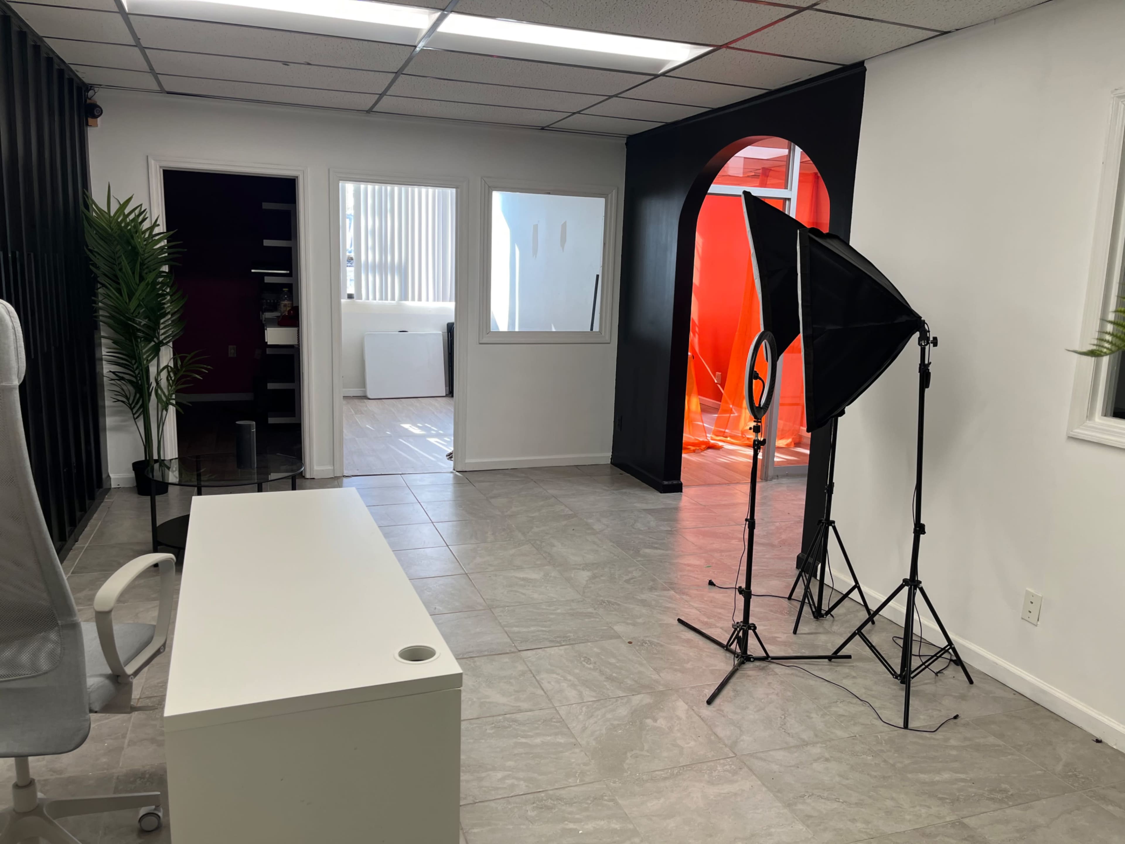 A spacious office interior with a desk, a chair, and a lighting setup in the corner, surrounded by white walls and a vibrant red archway.