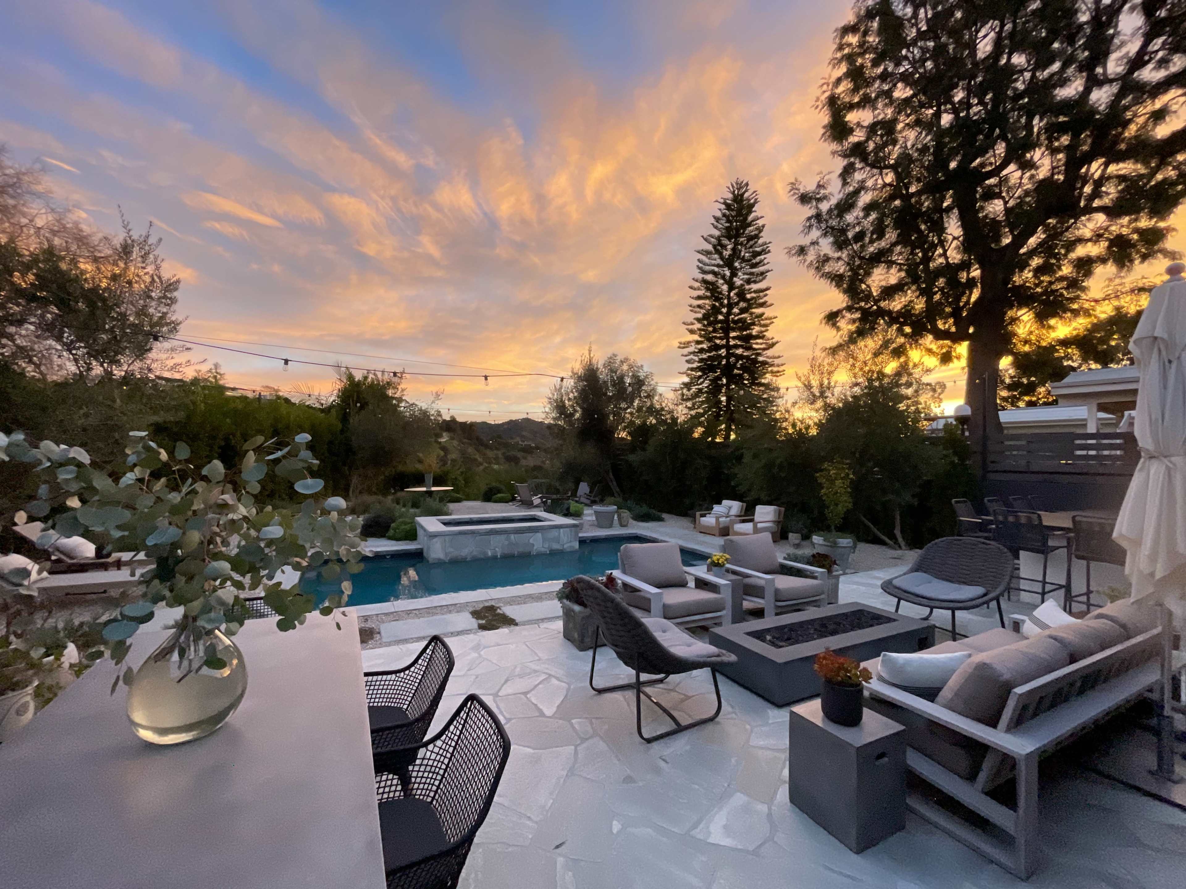 The image shows a backyard patio with a pool, seating areas, and a beautiful sunset sky filled with clouds.
