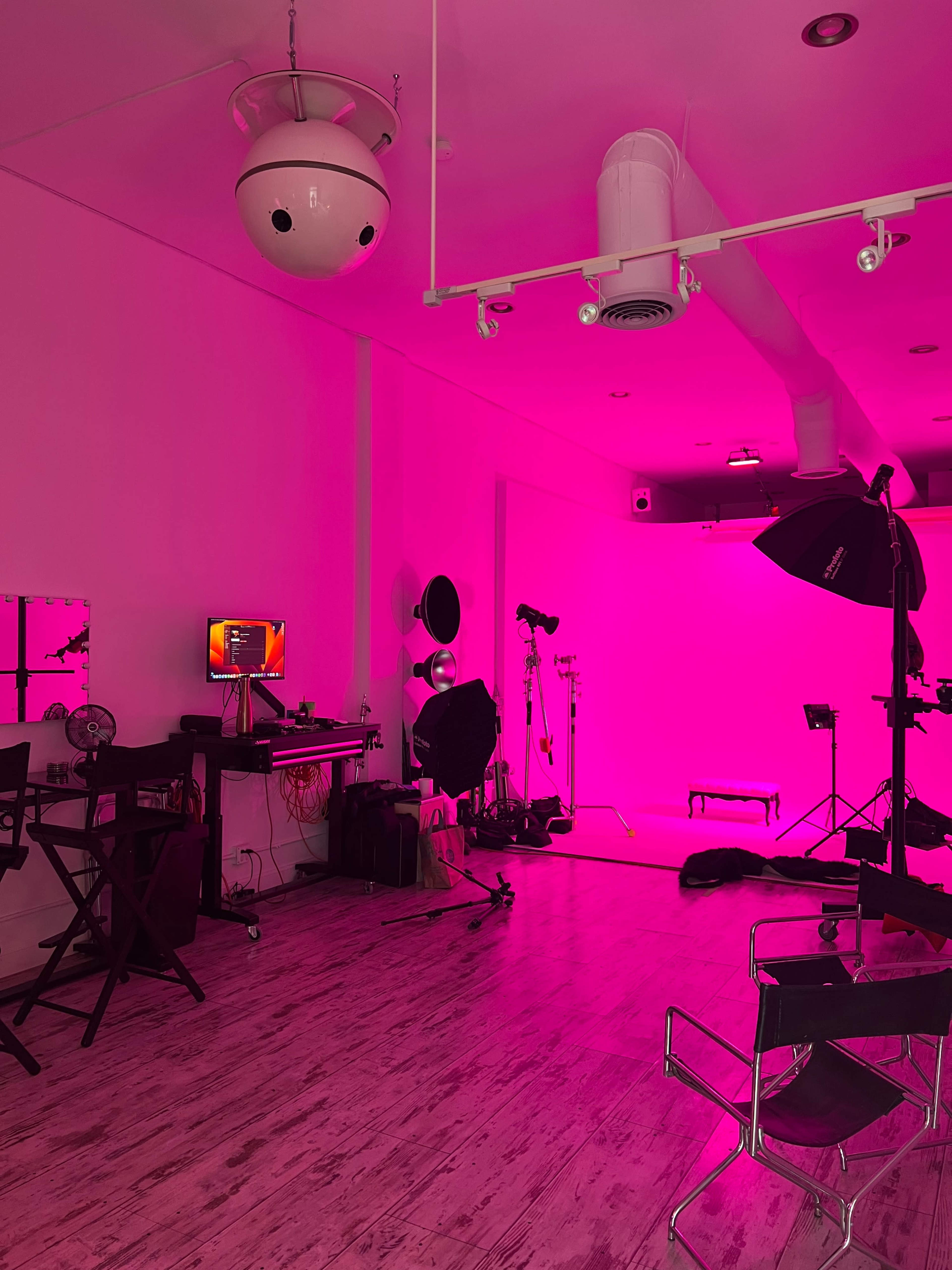 A photography studio is illuminated with pink lighting, featuring various equipment including softboxes, a backdrop, and a computer setup.