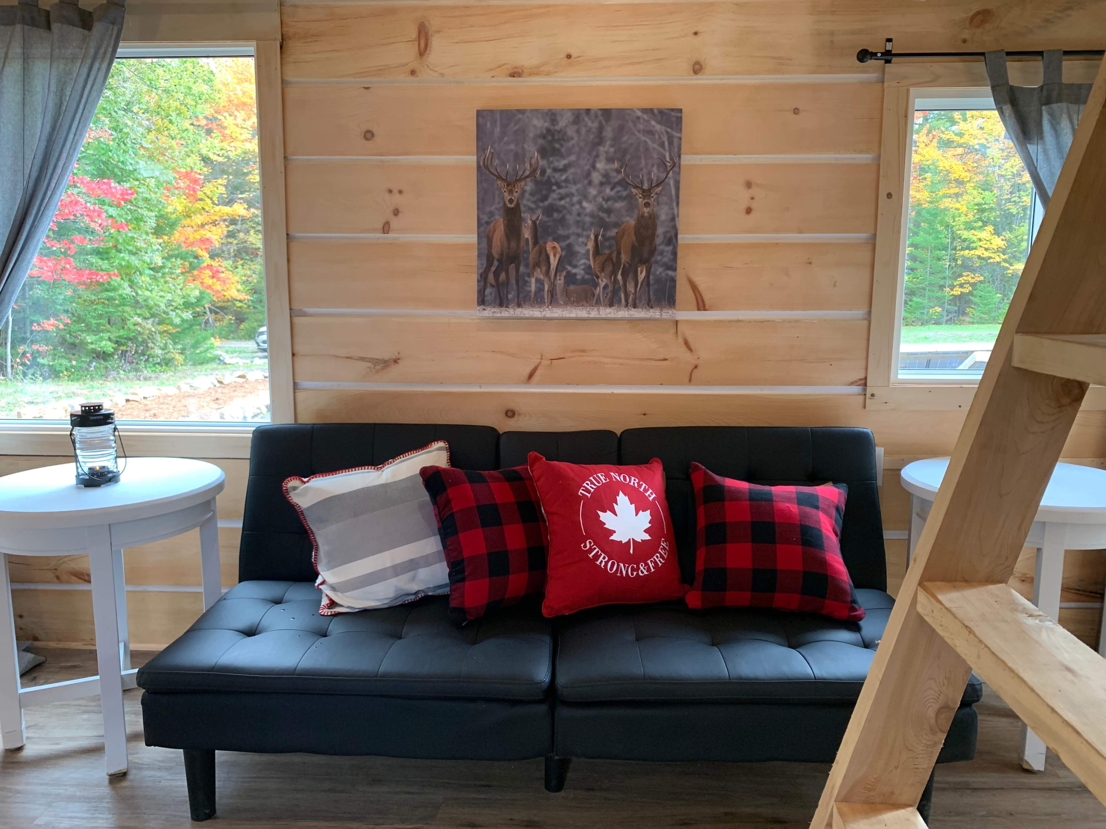 The image shows a cozy cabin interior with a black futon couch adorned with red and black checkered pillows, flanked by two small white tables and wooden walls.