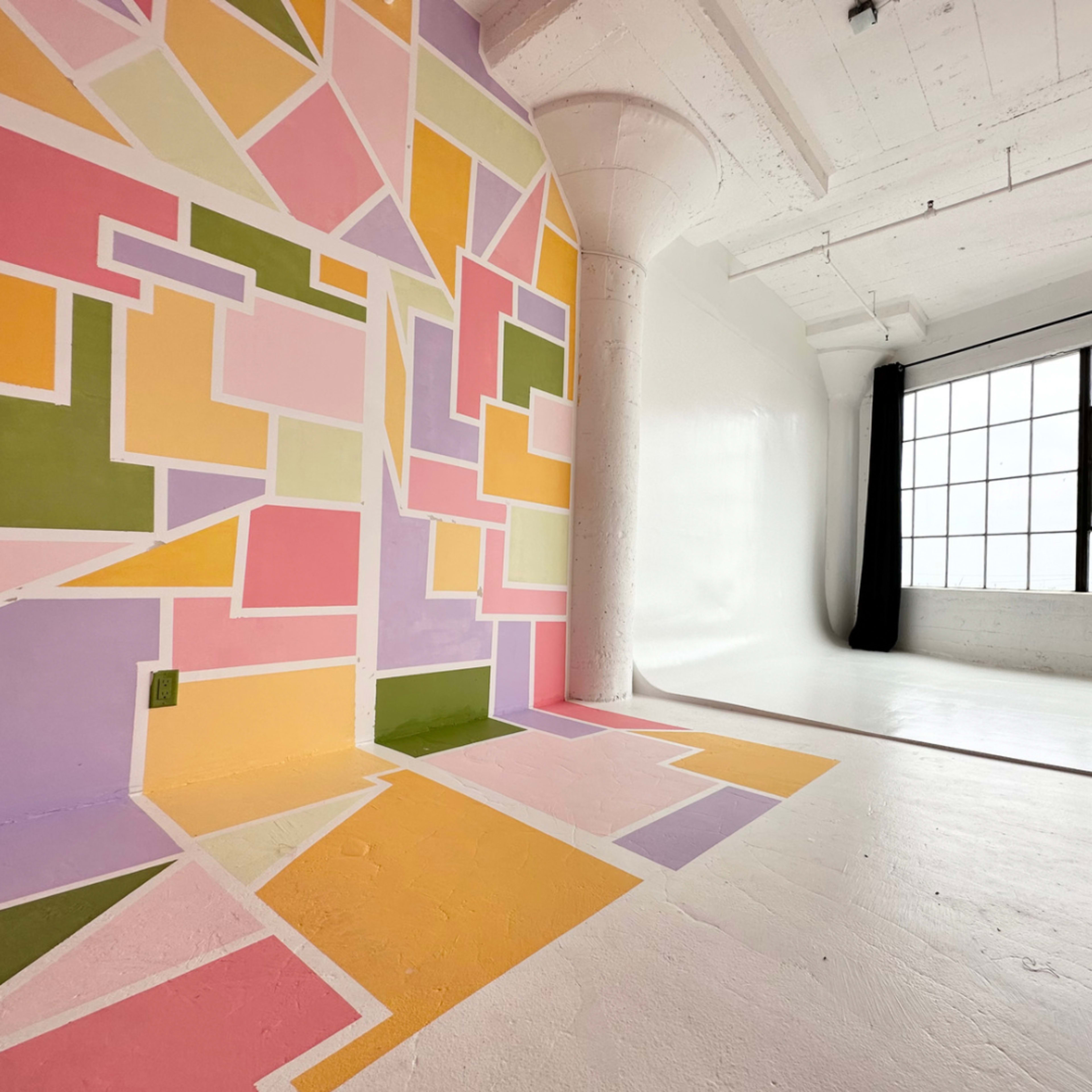 A studio space featuring a colorful geometric mural on one wall, with a large window and a plain white wall opposite.