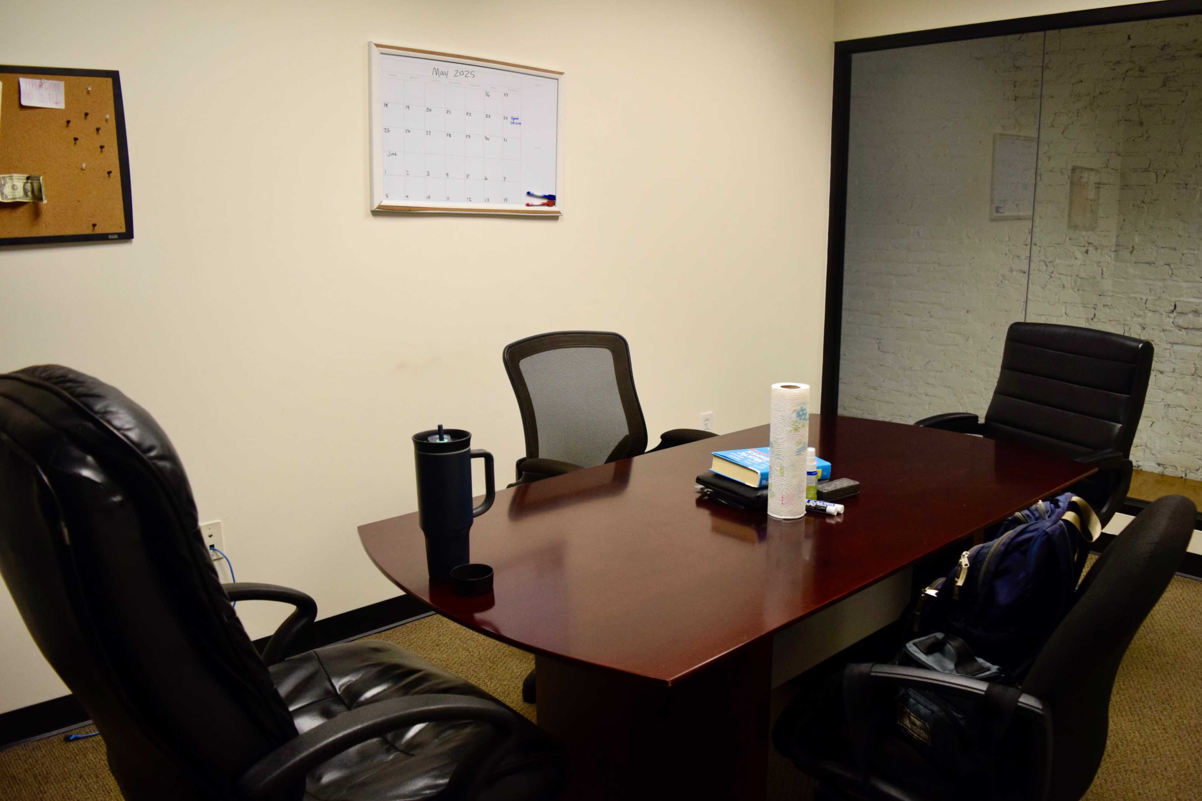 A conference room is shown with a brown table surrounded by four black chairs, and a calendar hangs on the wall.