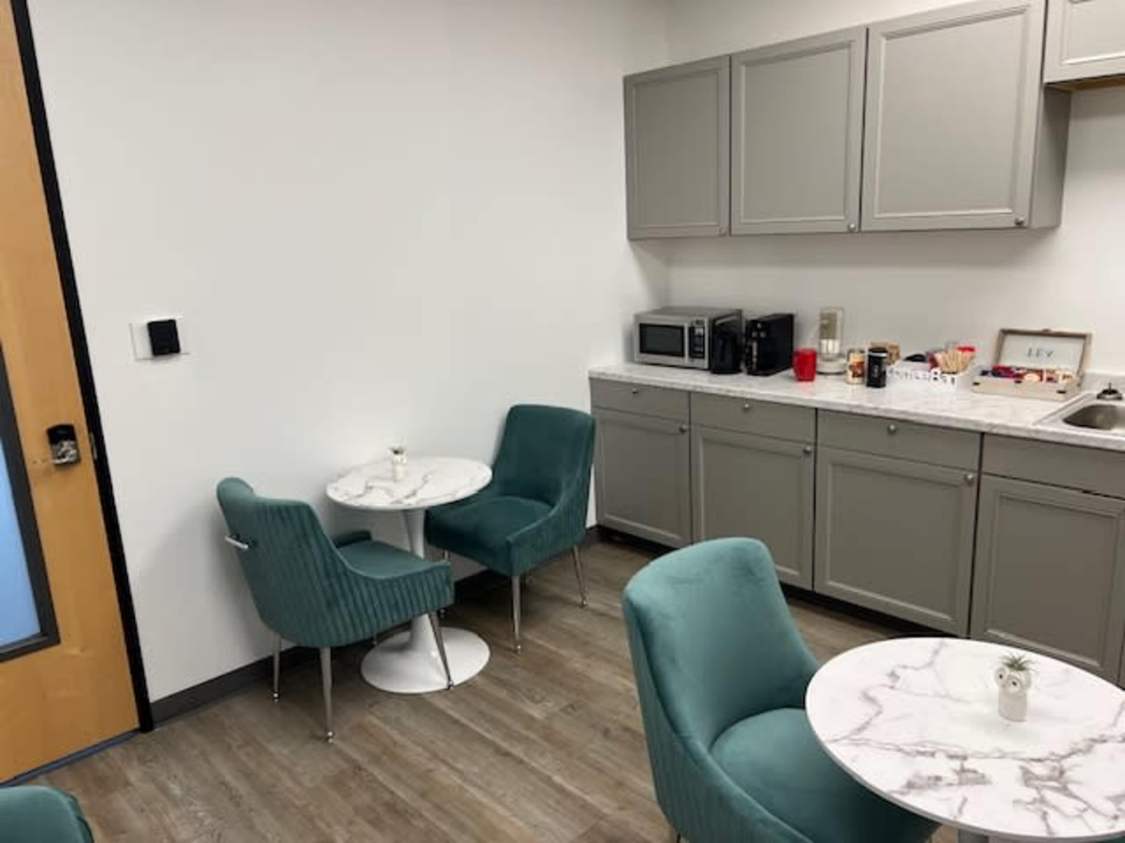 The image shows a small break room with two round tables and teal chairs, along with kitchen cabinets, a microwave, and various snacks on the countertop.