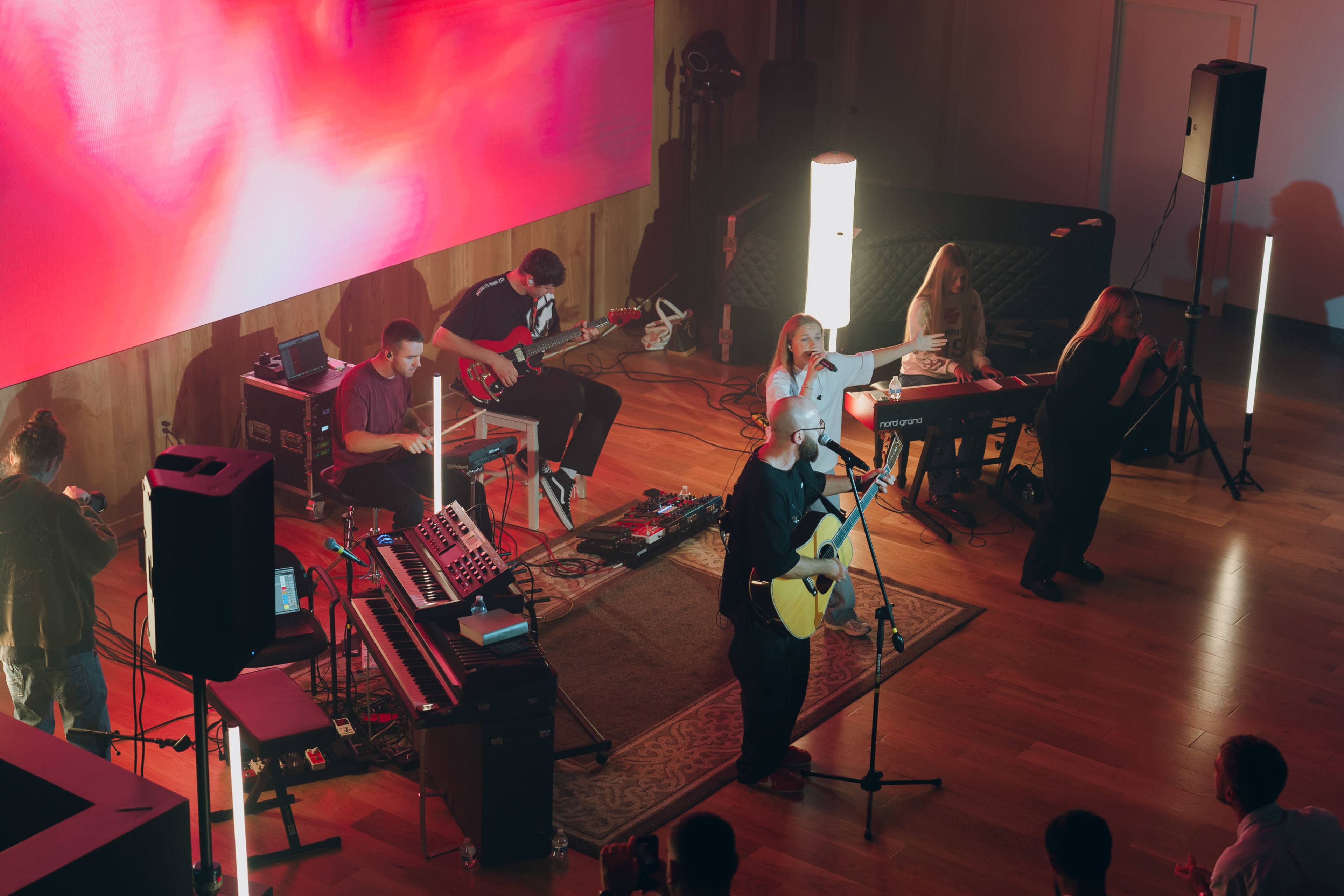 A band performs on stage, with a vocalist and musicians playing instruments in front of a large, colorful light display.
