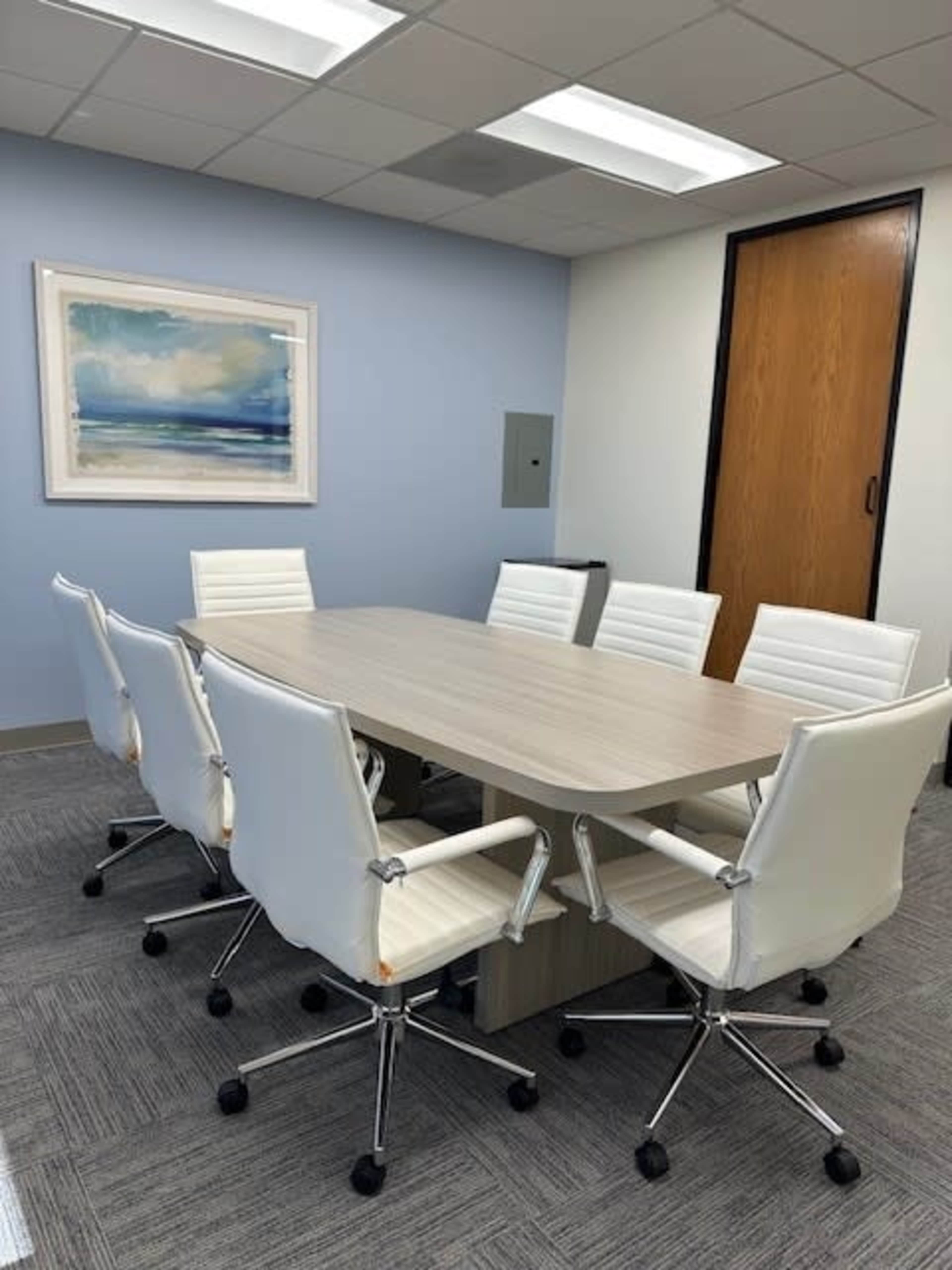 A modern conference room features a light gray table surrounded by six white ergonomic chairs, with a painting hanging on the wall.