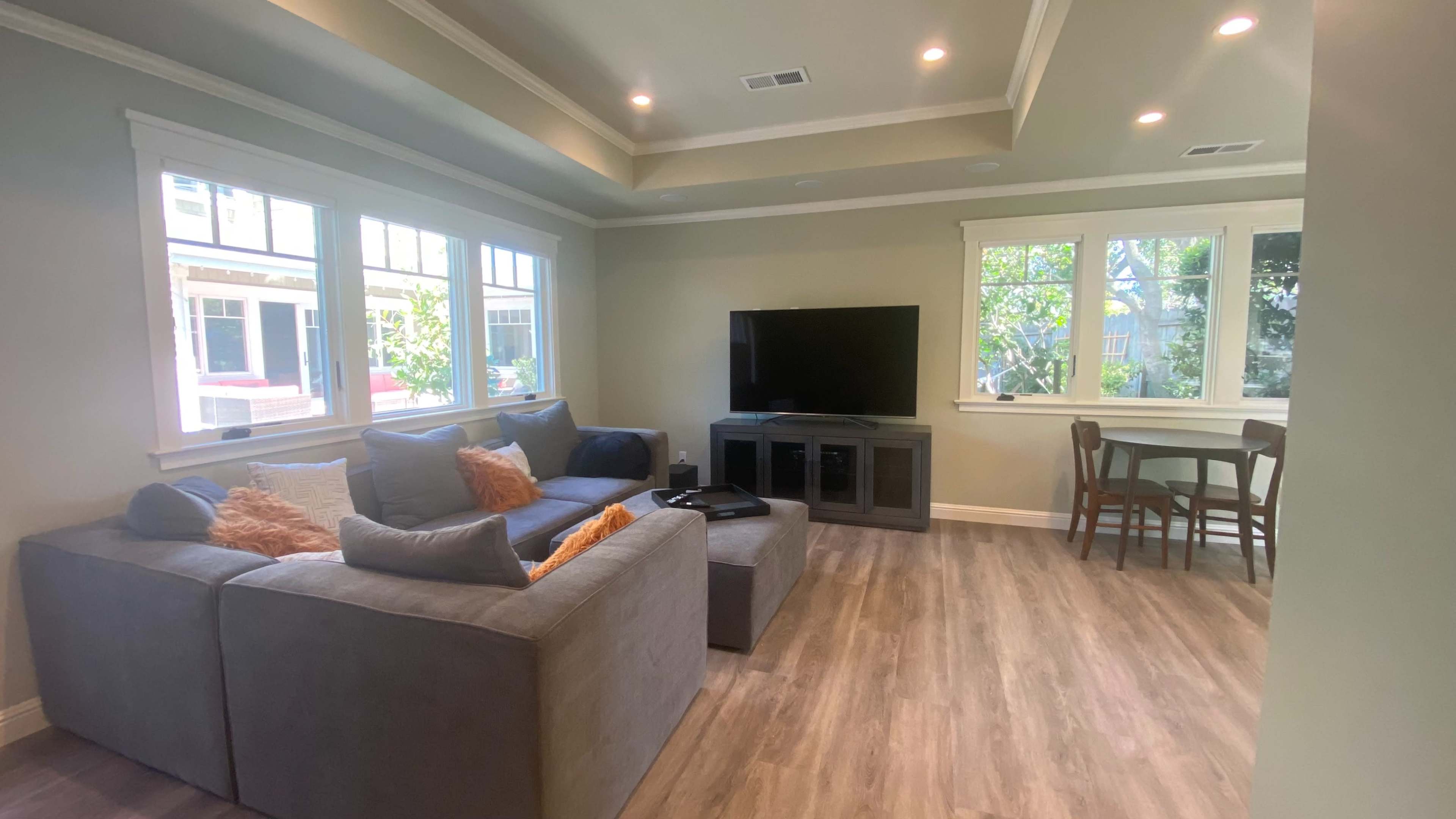 The living room features a gray sectional sofa, a television on a stand, and a dining area with a table and chairs, all illuminated by natural light from multiple windows.