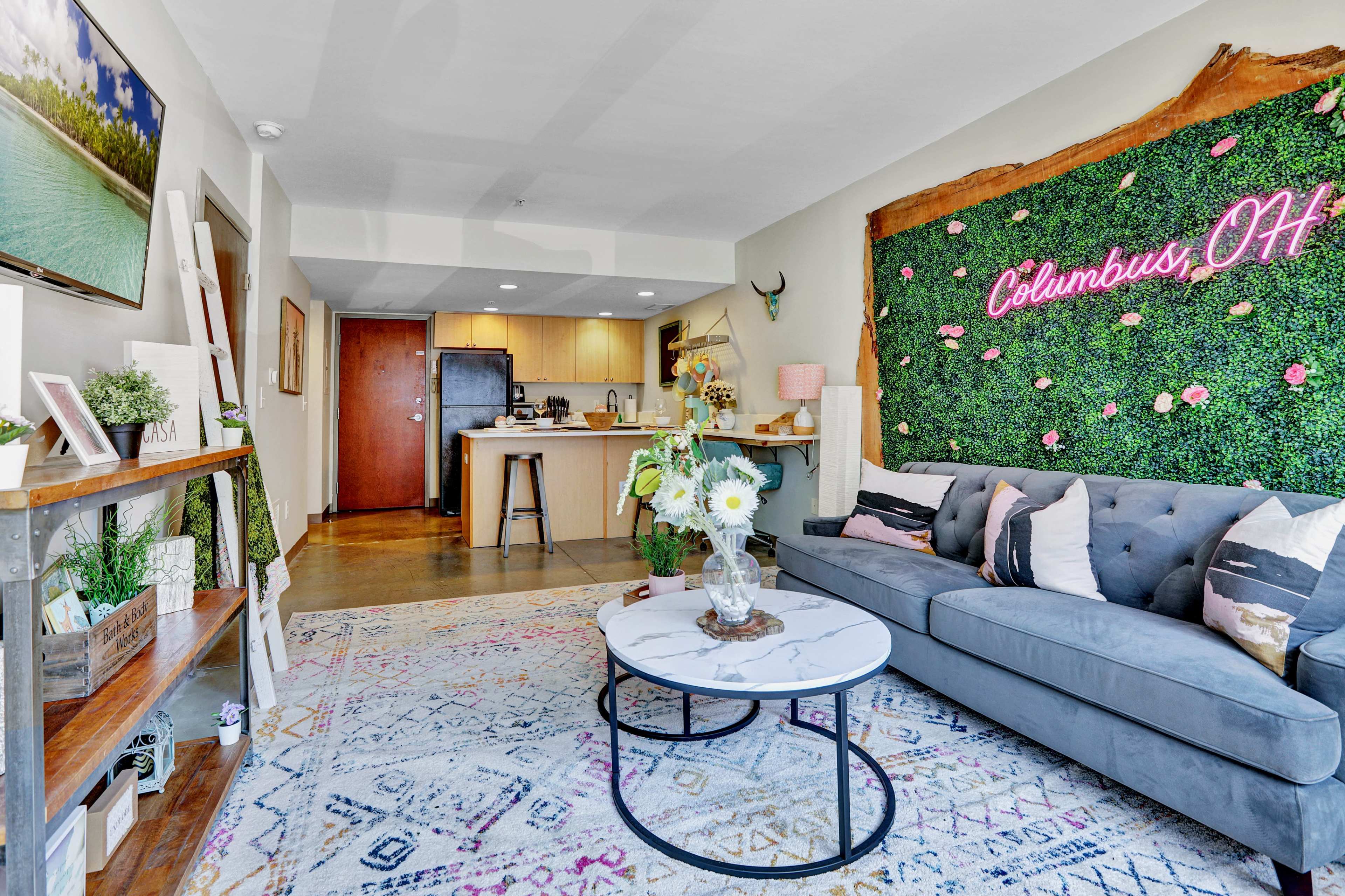 A living room with a gray sofa, a round coffee table, a decorative green wall featuring the text "Columbus, OH," and an open kitchen in the background.