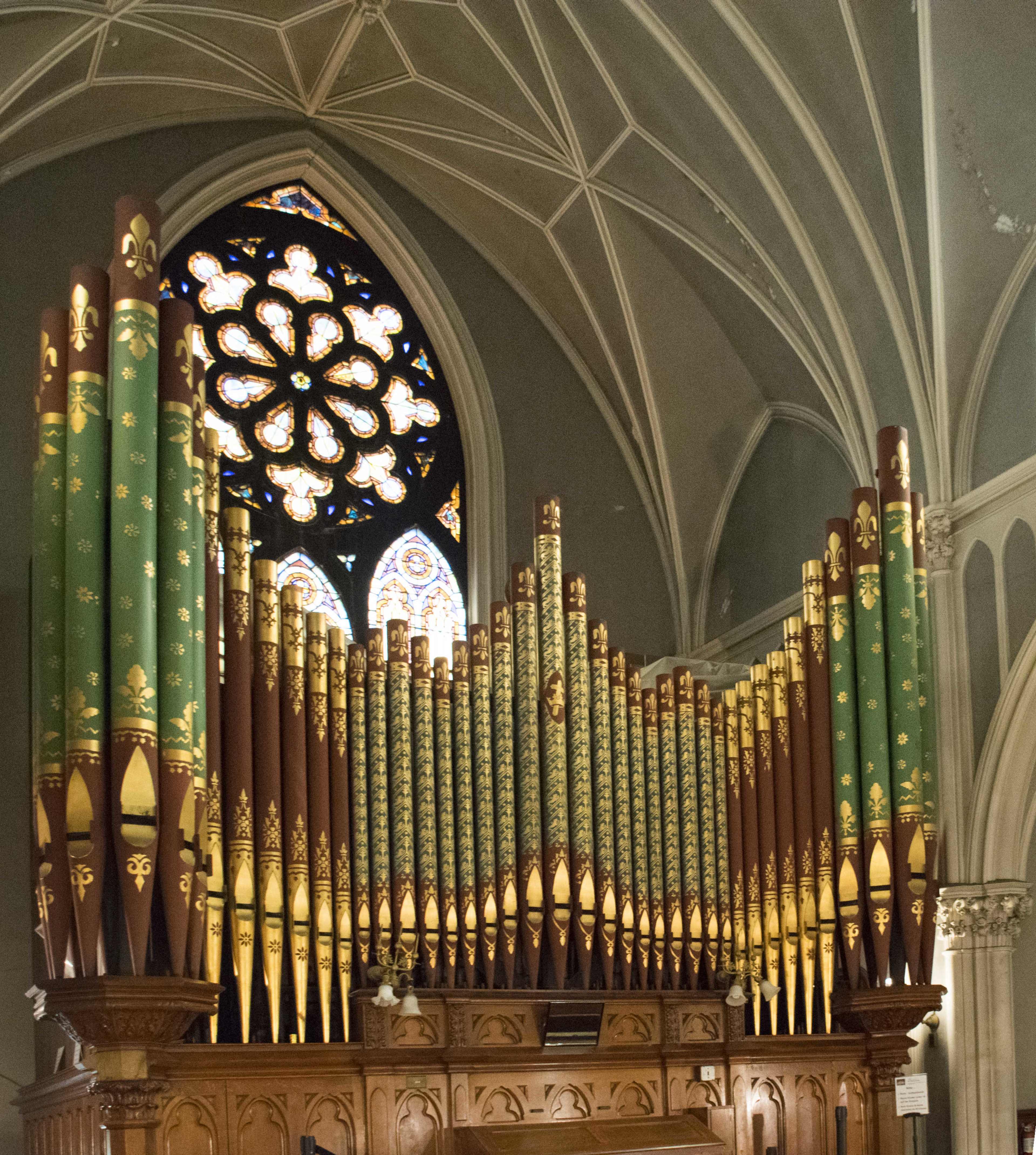 The image shows an ornate pipe organ inside a cathedral, featuring tall, multi-colored pipes and a stained glass window in the background.