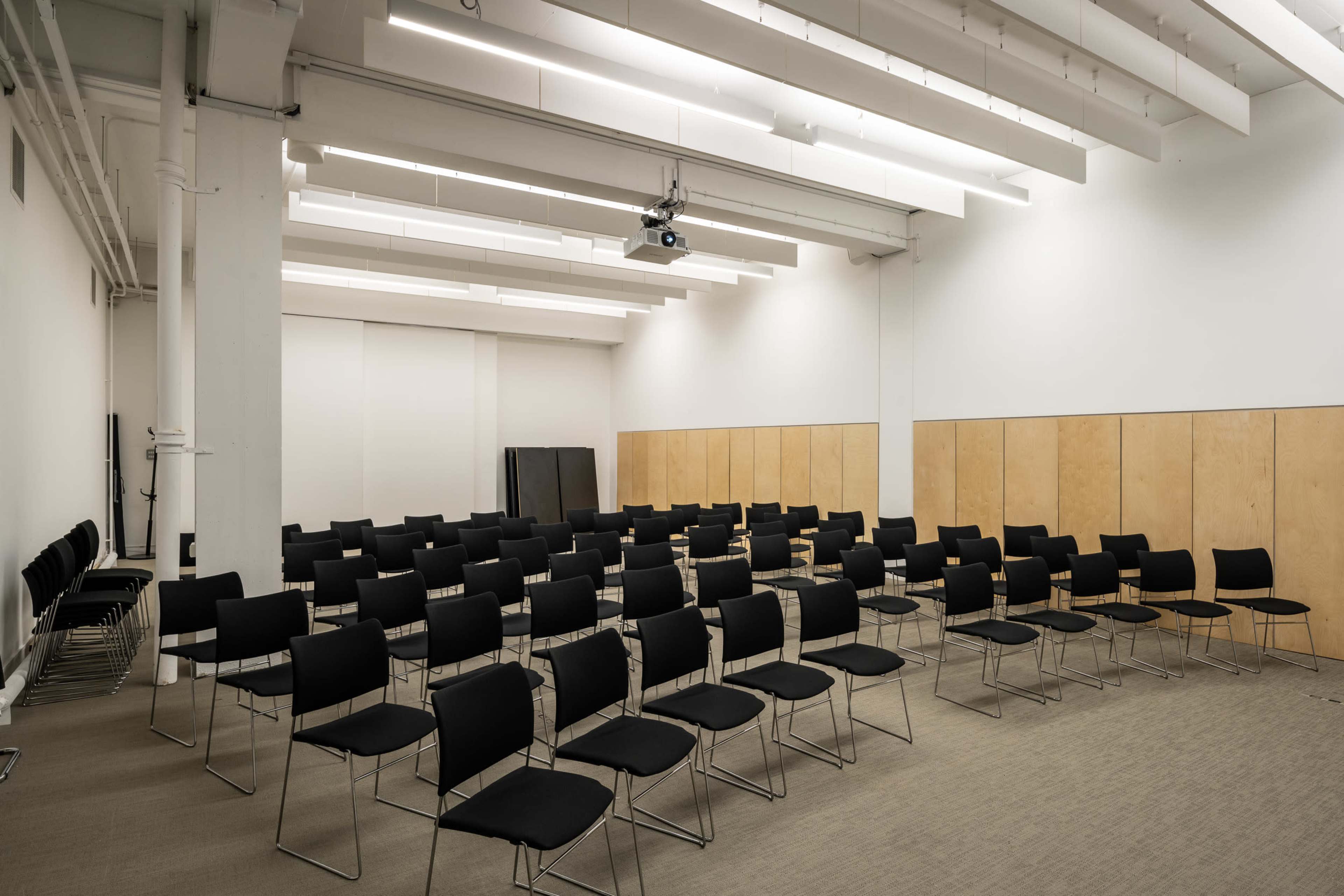 The image shows a large, empty room with a grid of black chairs arranged in rows facing a blank wall, illuminated by overhead lighting and featuring wooden paneling.