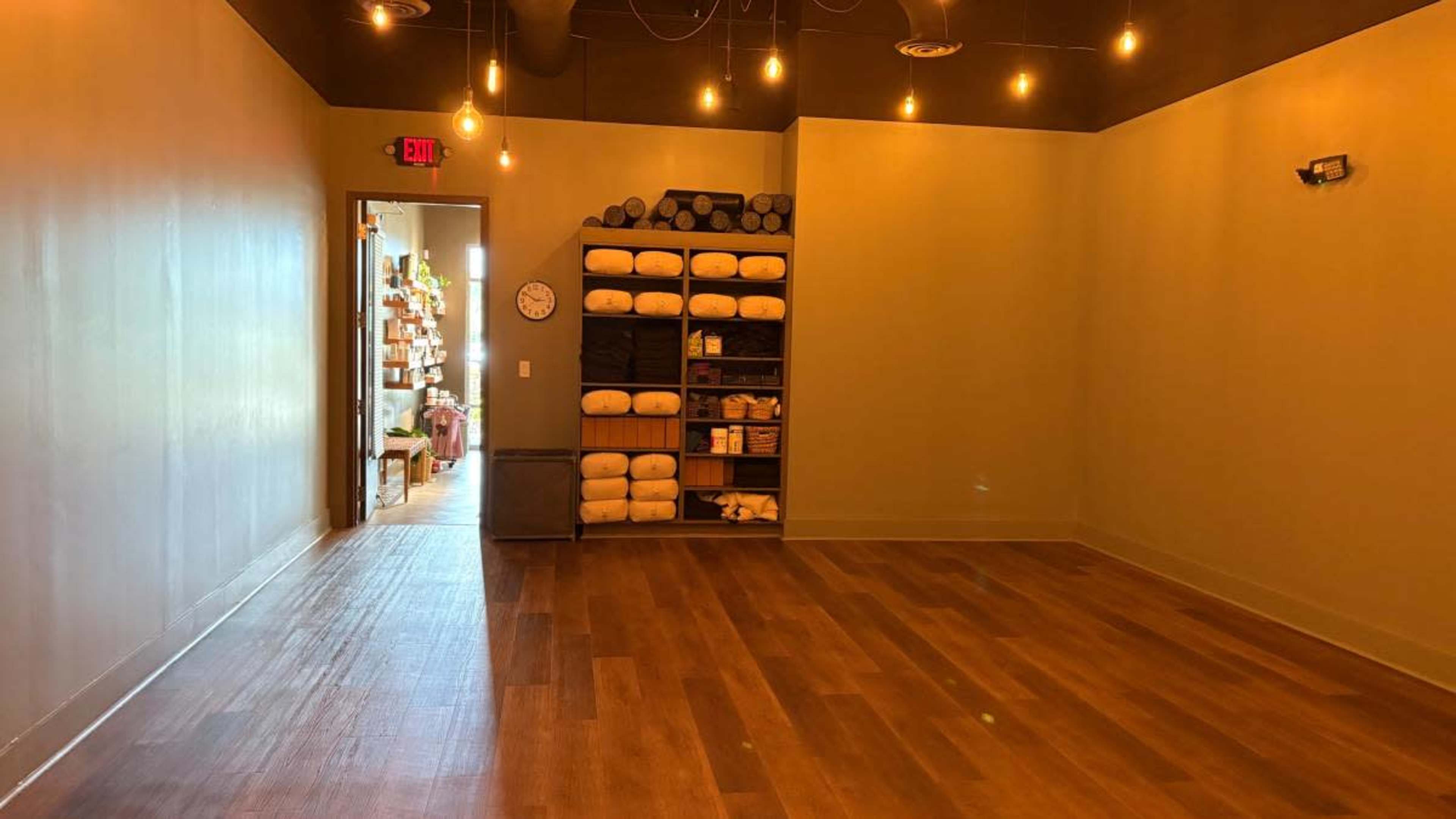 The image shows a spacious, minimalist room with wooden flooring, a shelf stacked with rolled yoga mats, and warm overhead lighting.