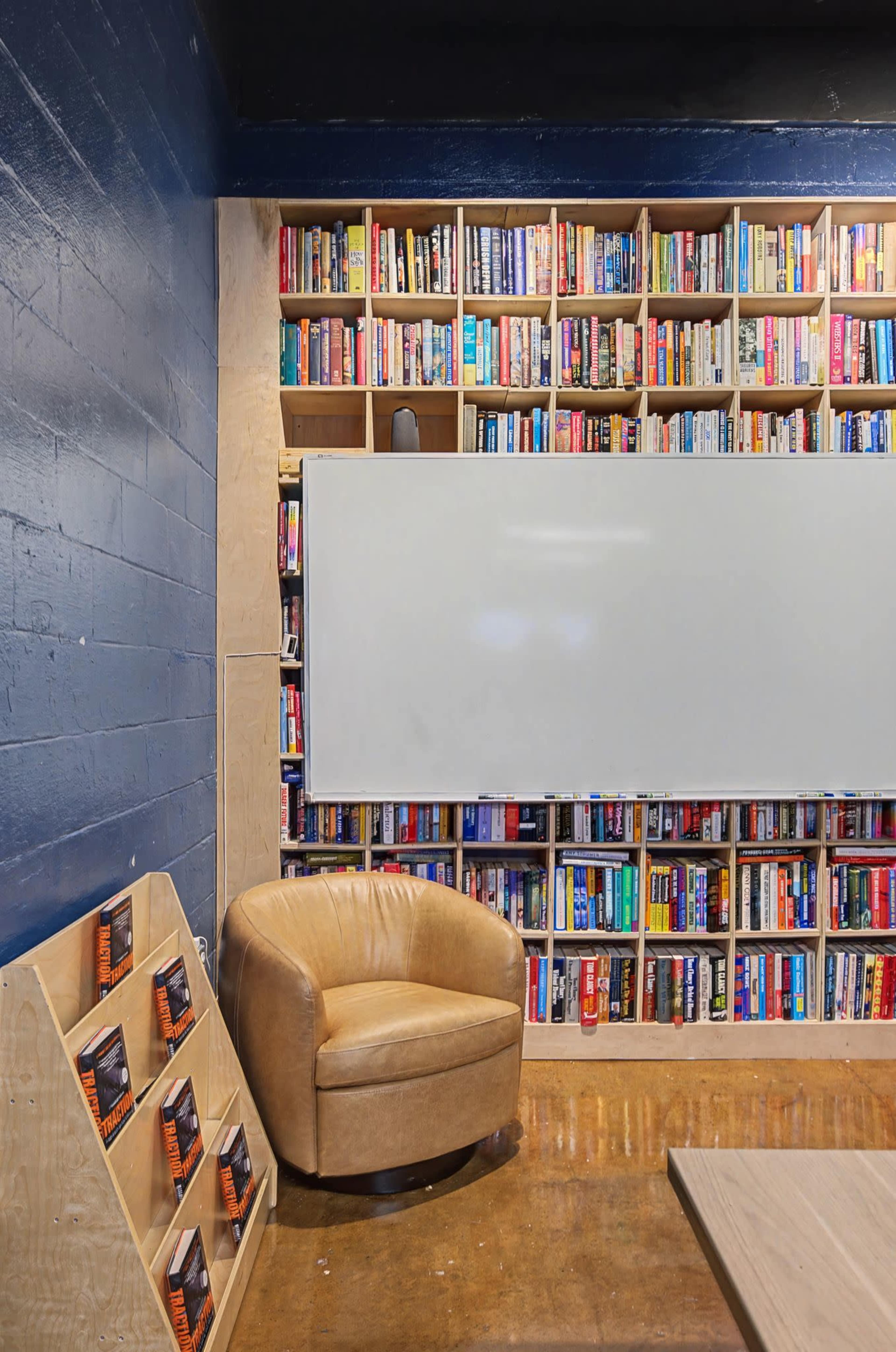 A cozy reading nook features a brown leather chair next to a wall filled with bookshelves and a whiteboard.