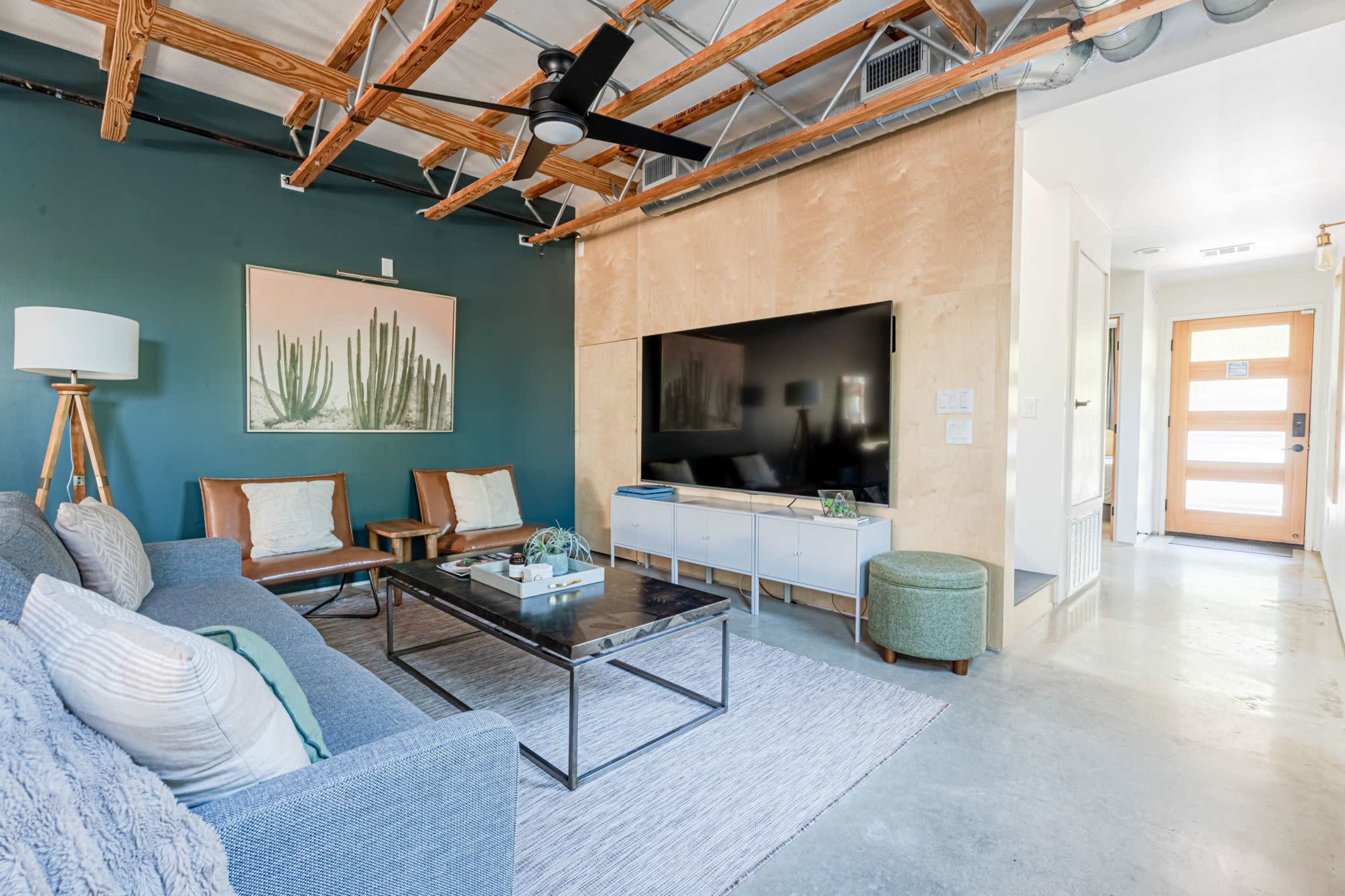 The living room features a modern design with a gray sofa, wooden ceiling beams, a large television mounted on a wall, and a combination of decorative furniture and artwork.