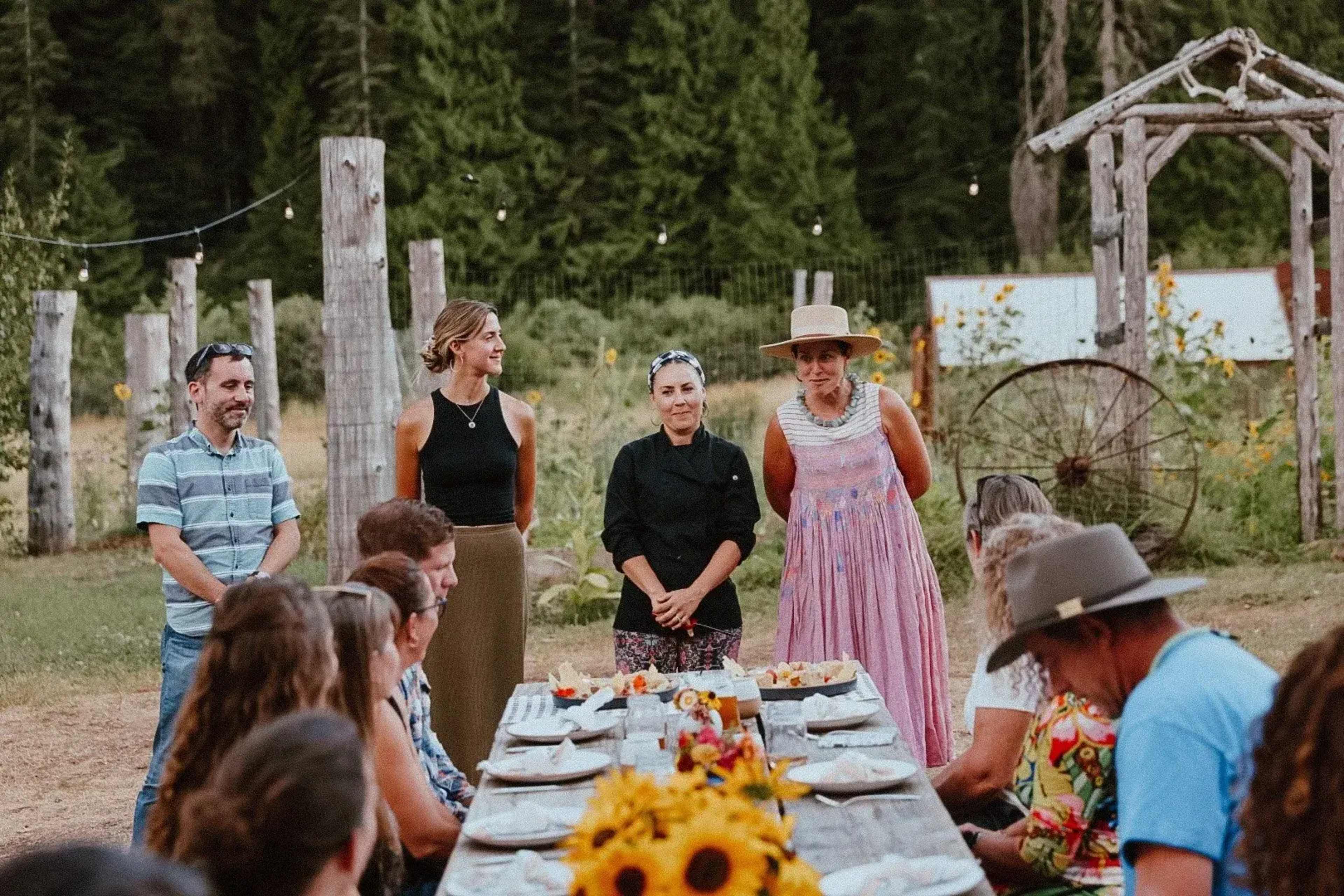 A group of people stands around a long outdoor table set for a meal in a rustic, natural setting.