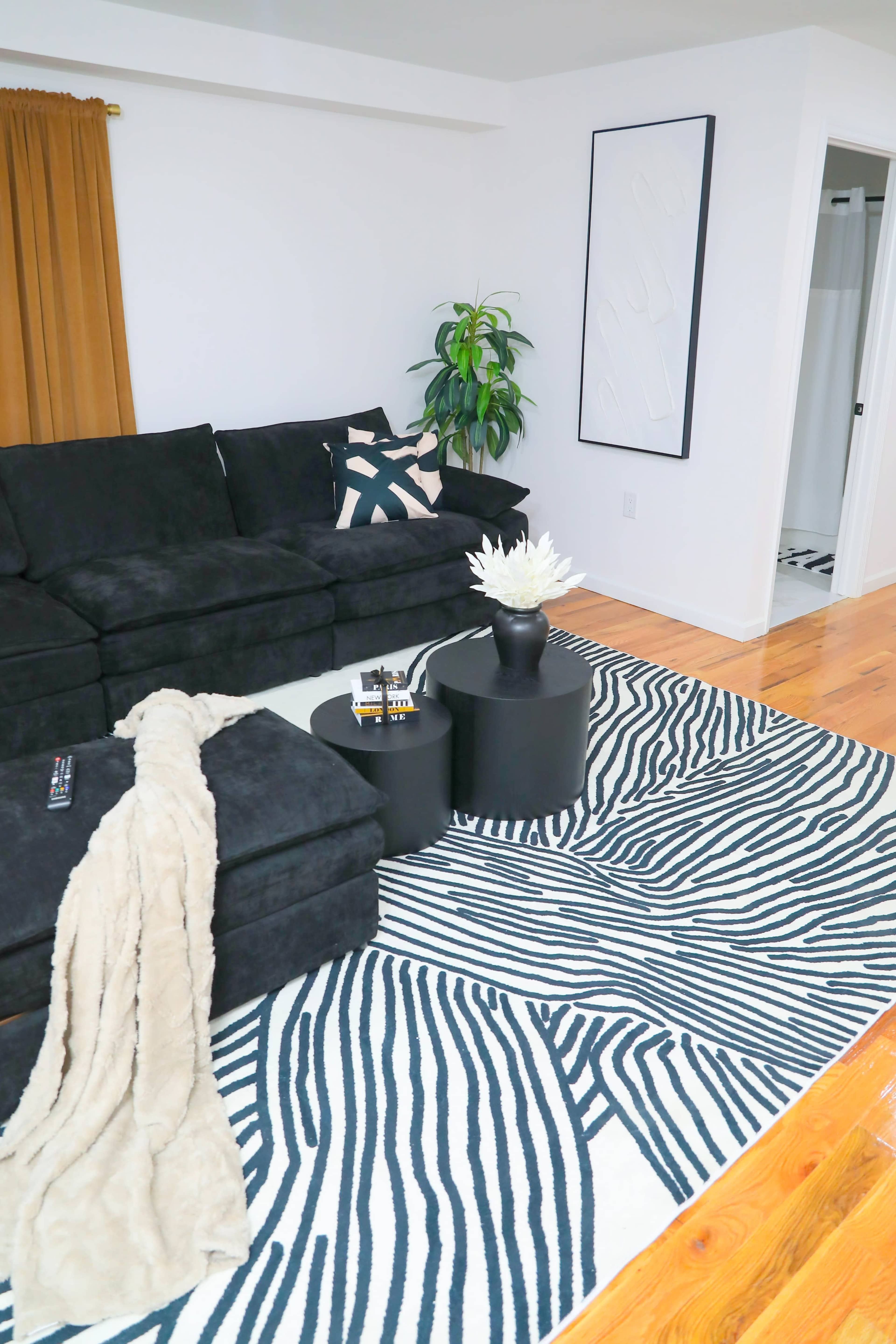 The living room features a black sectional sofa, a patterned area rug, two black side tables, and a potted plant in a bright space with wooden flooring.