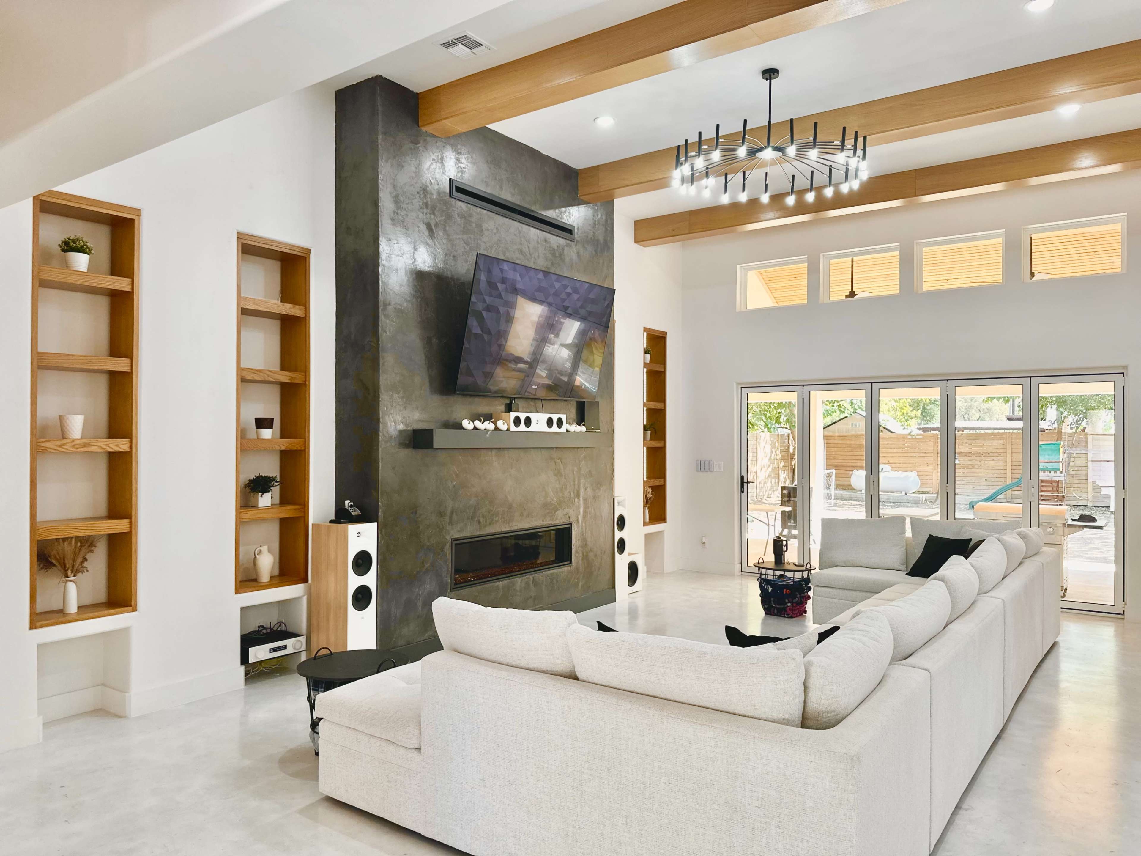 The image shows a modern living room featuring a large sectional sofa, a mounted television above a sleek fireplace, and built-in shelves on either side of a textured accent wall.