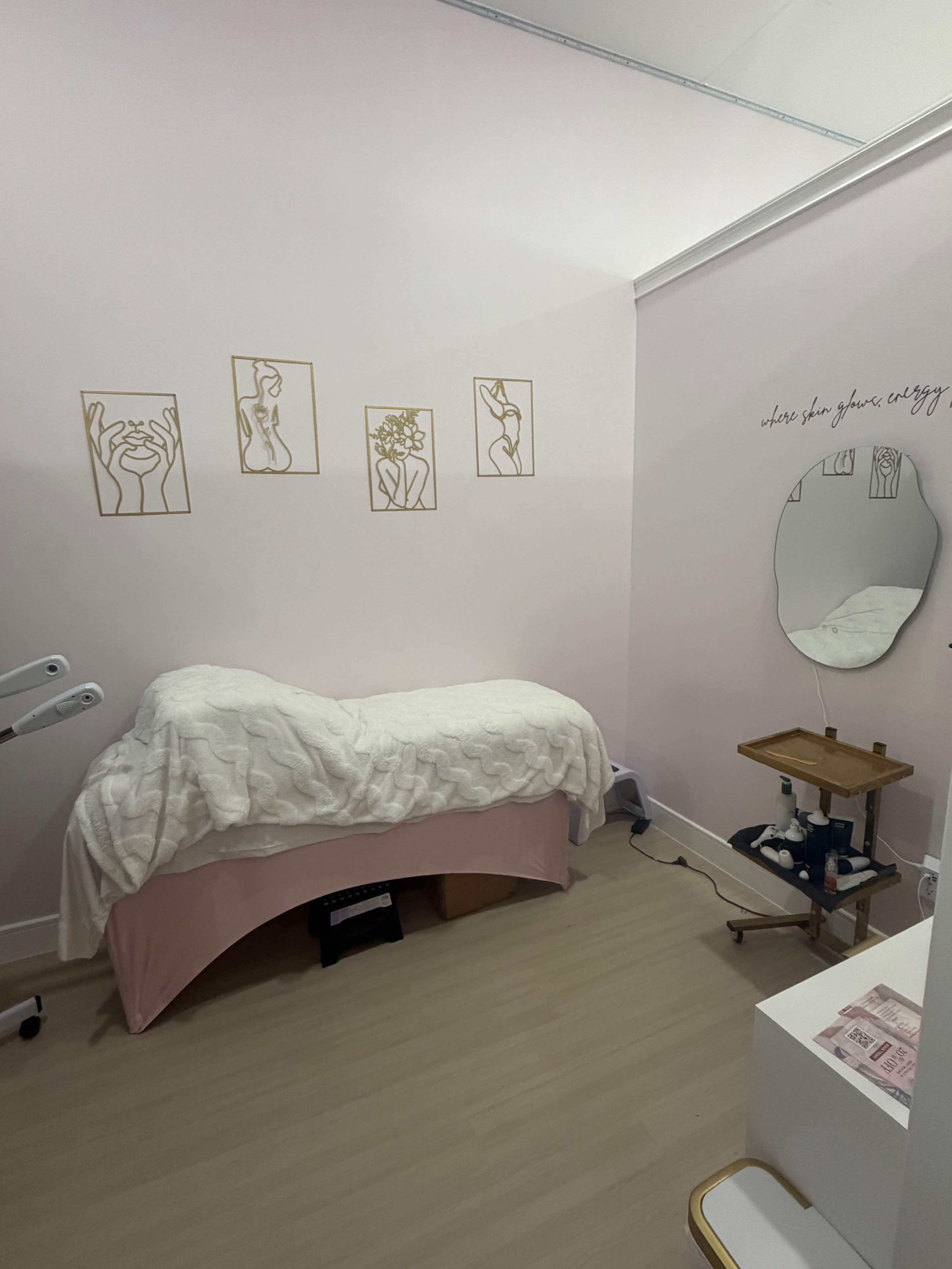 The image shows a treatment room with a bed covered in a white quilt and light pink skirt, adorned with wall art depicting minimalistic line drawings of hands and flowers, and a mirror positioned beside a small wooden table.