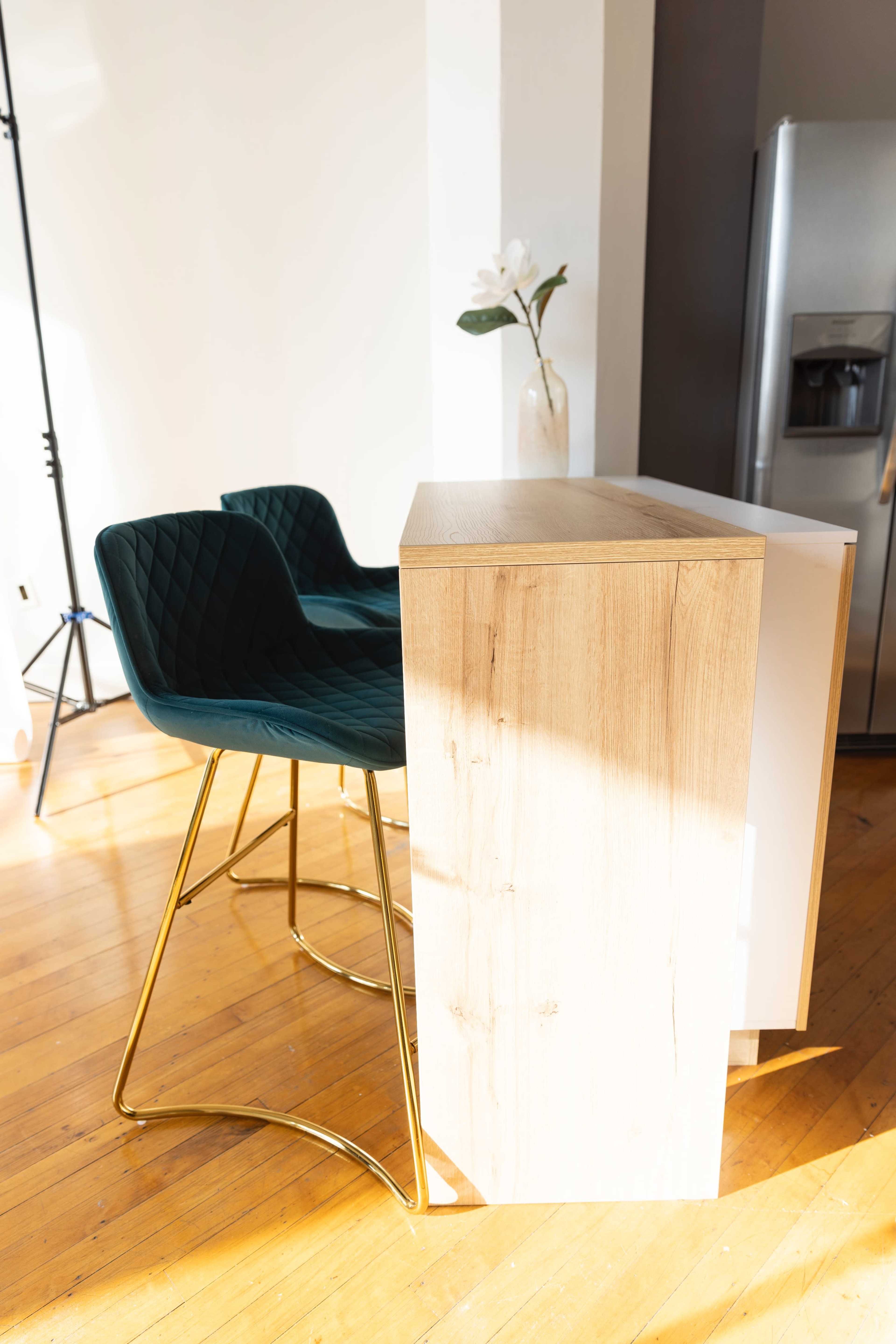 A wooden bar counter with a light finish stands next to two green velvet bar stools on a hardwood floor.