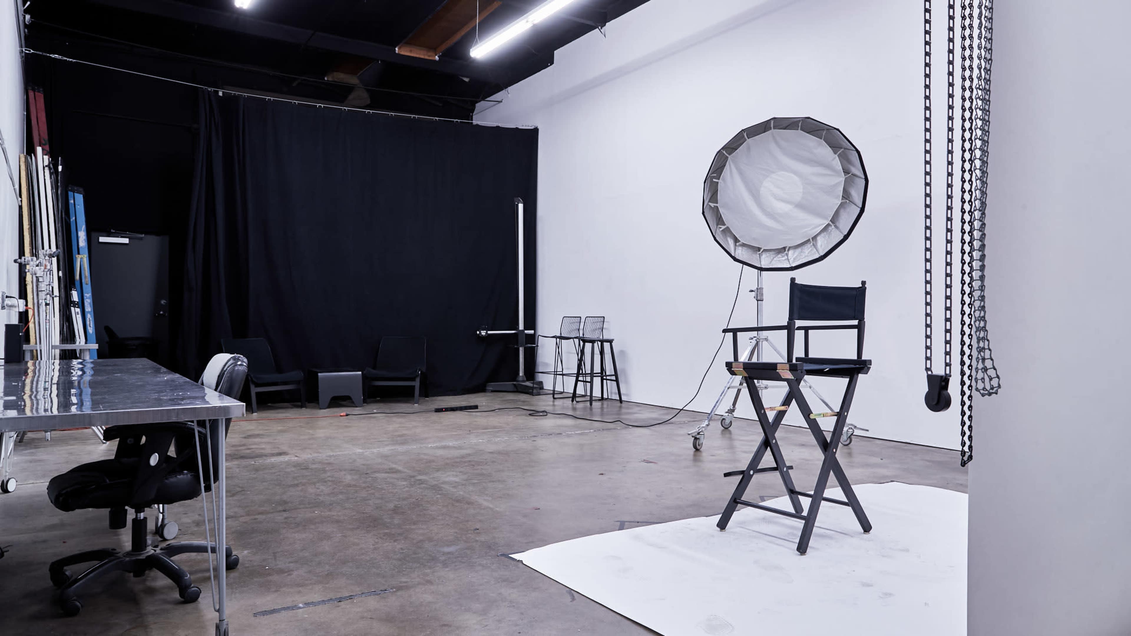 A photo studio features a director's chair on a white backdrop, with lighting equipment and a black curtain in the background.