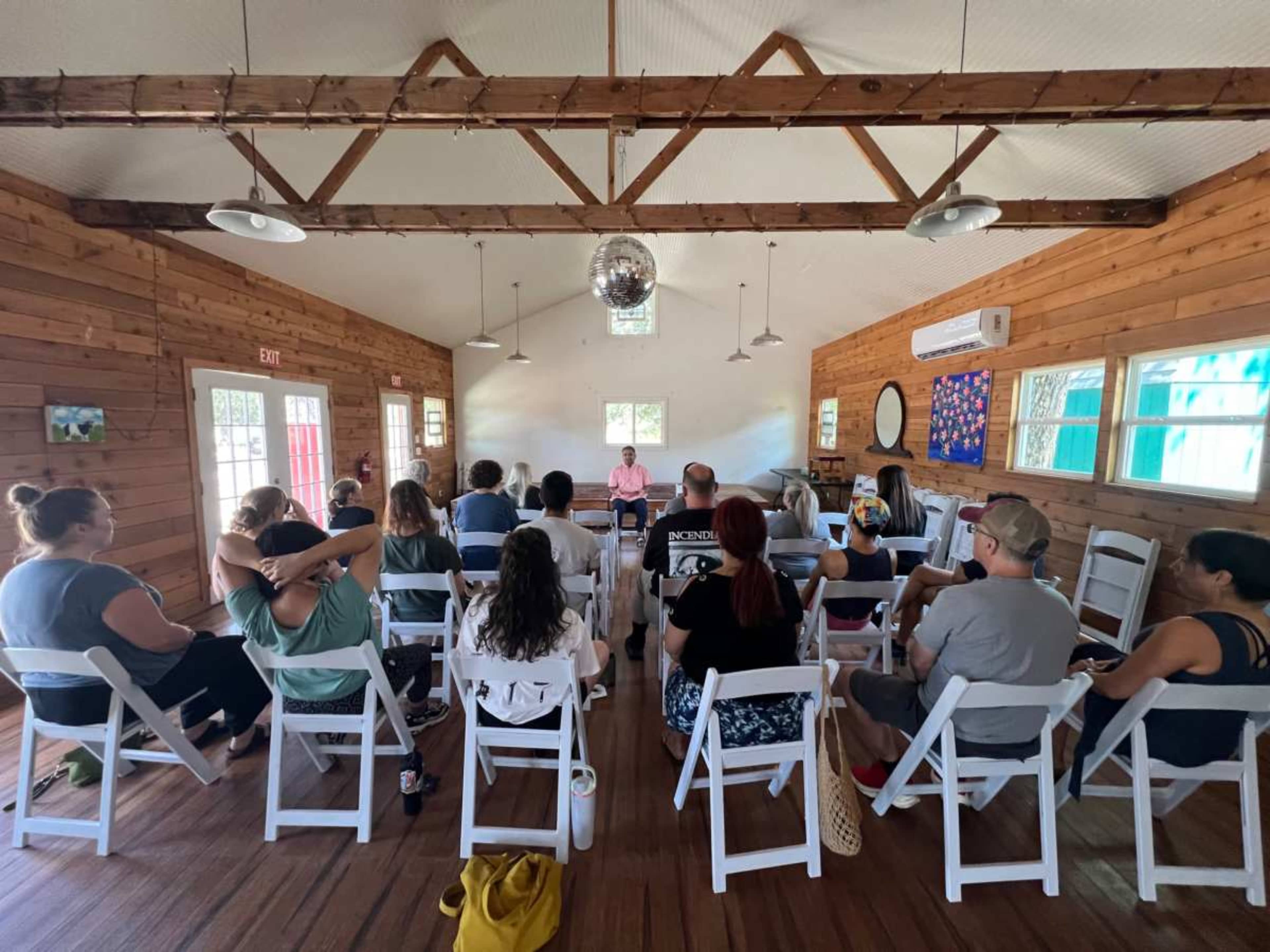 A group of people is seated in white chairs facing a speaker in a wooden interior room with large windows and a disco ball.