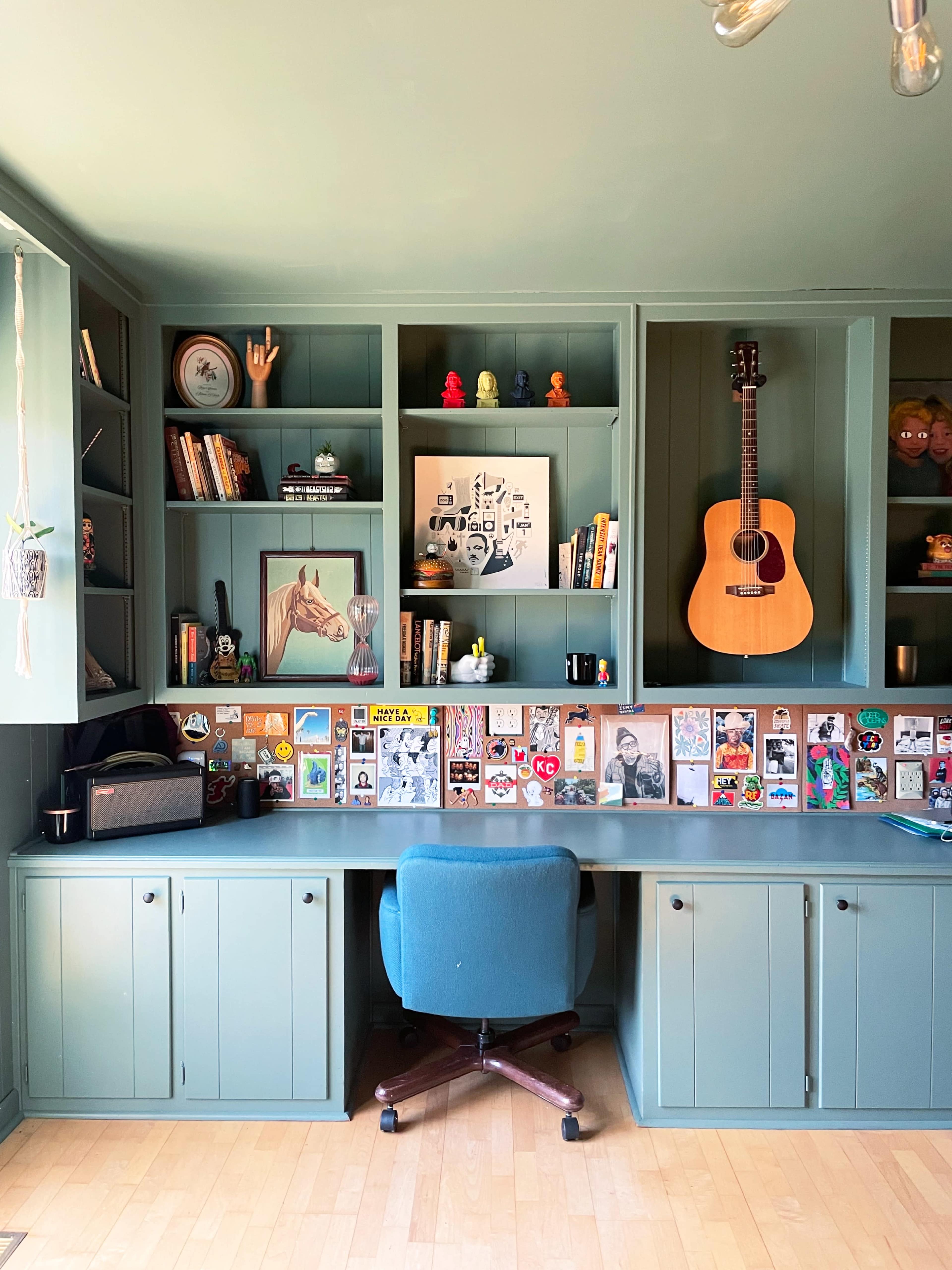 A home office features a mint green wall unit filled with books, decorations, and a guitar, with a rounded blue chair positioned at a spacious desk adorned with various framed pictures and stickers.