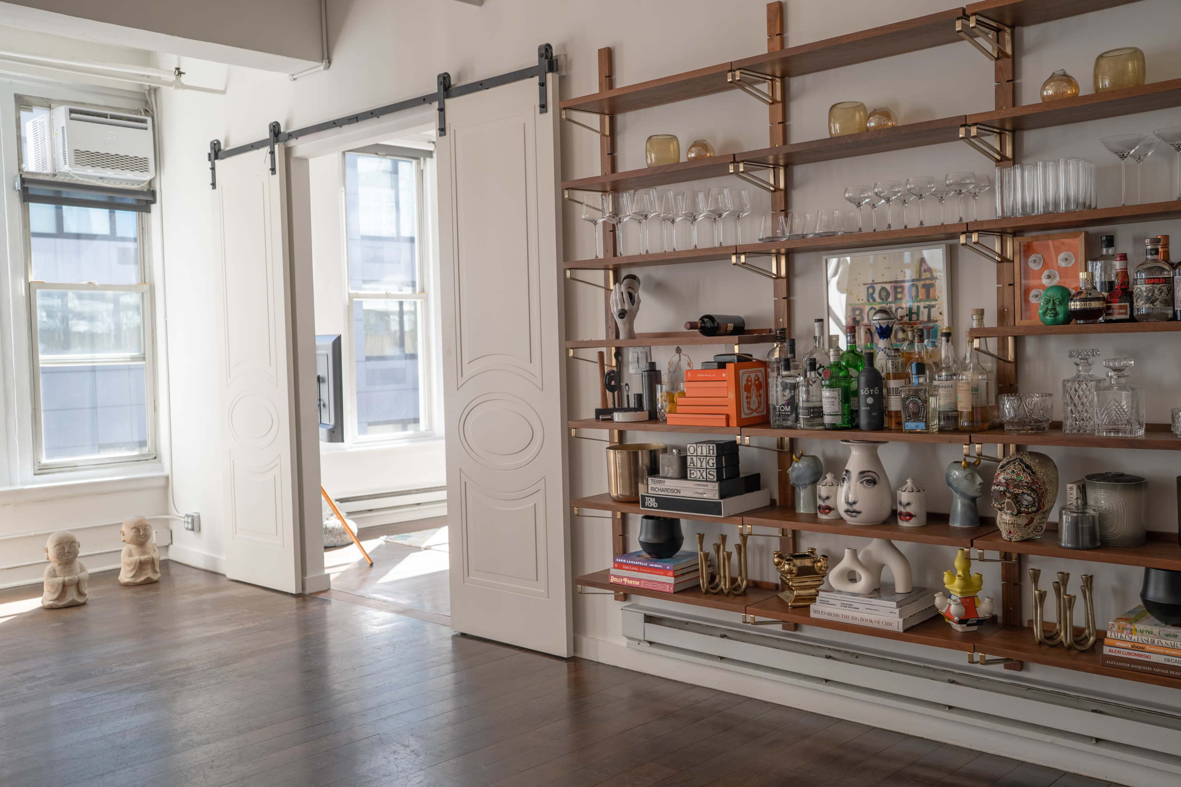 The image features a well-furnished room with large windows, a sliding door, and a decorative shelving unit displaying various glassware and decor items.