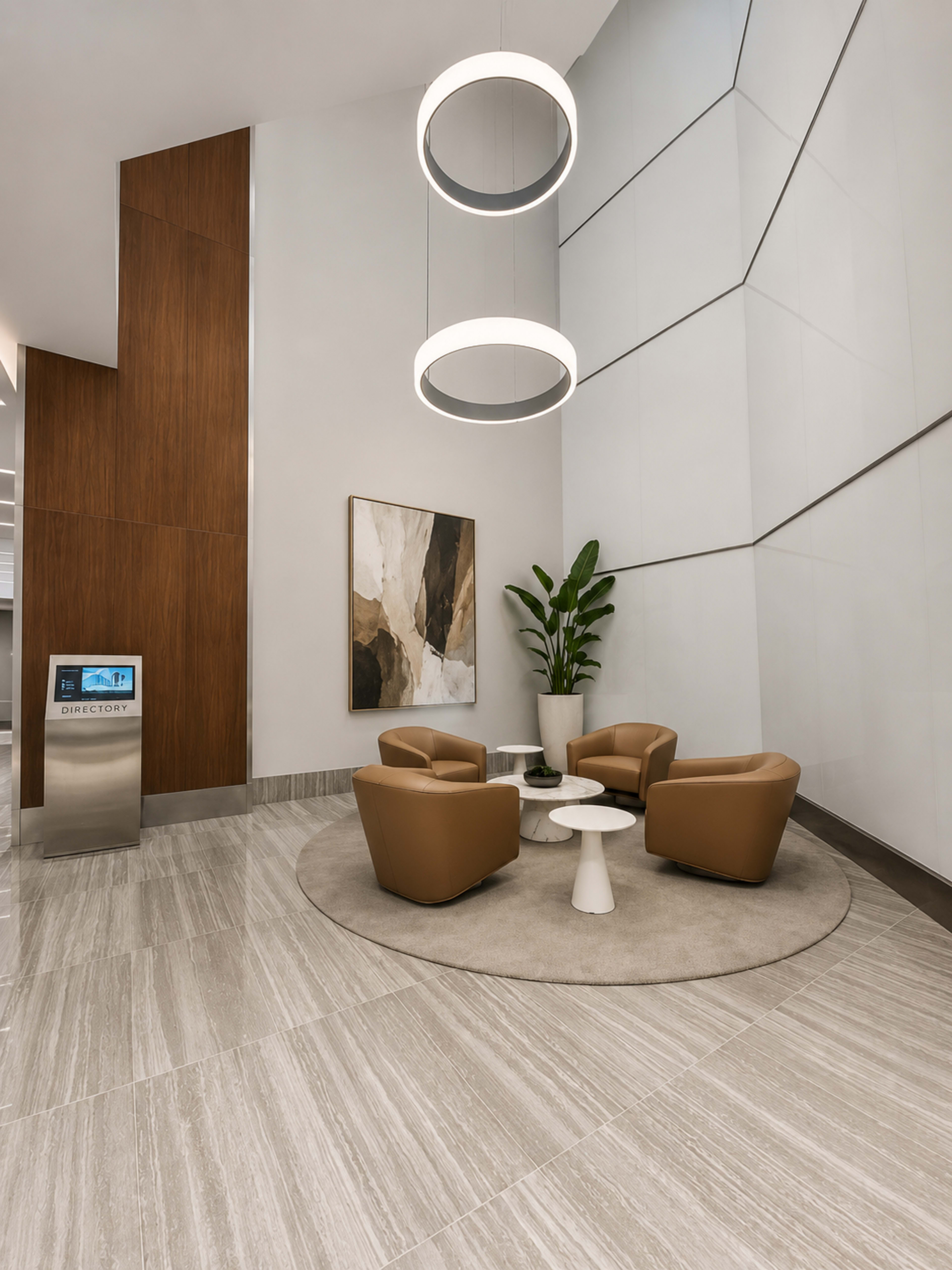 The image shows a contemporary lobby area with two round light fixtures, two brown armchairs, a small white table, and a large abstract painting on the wall.