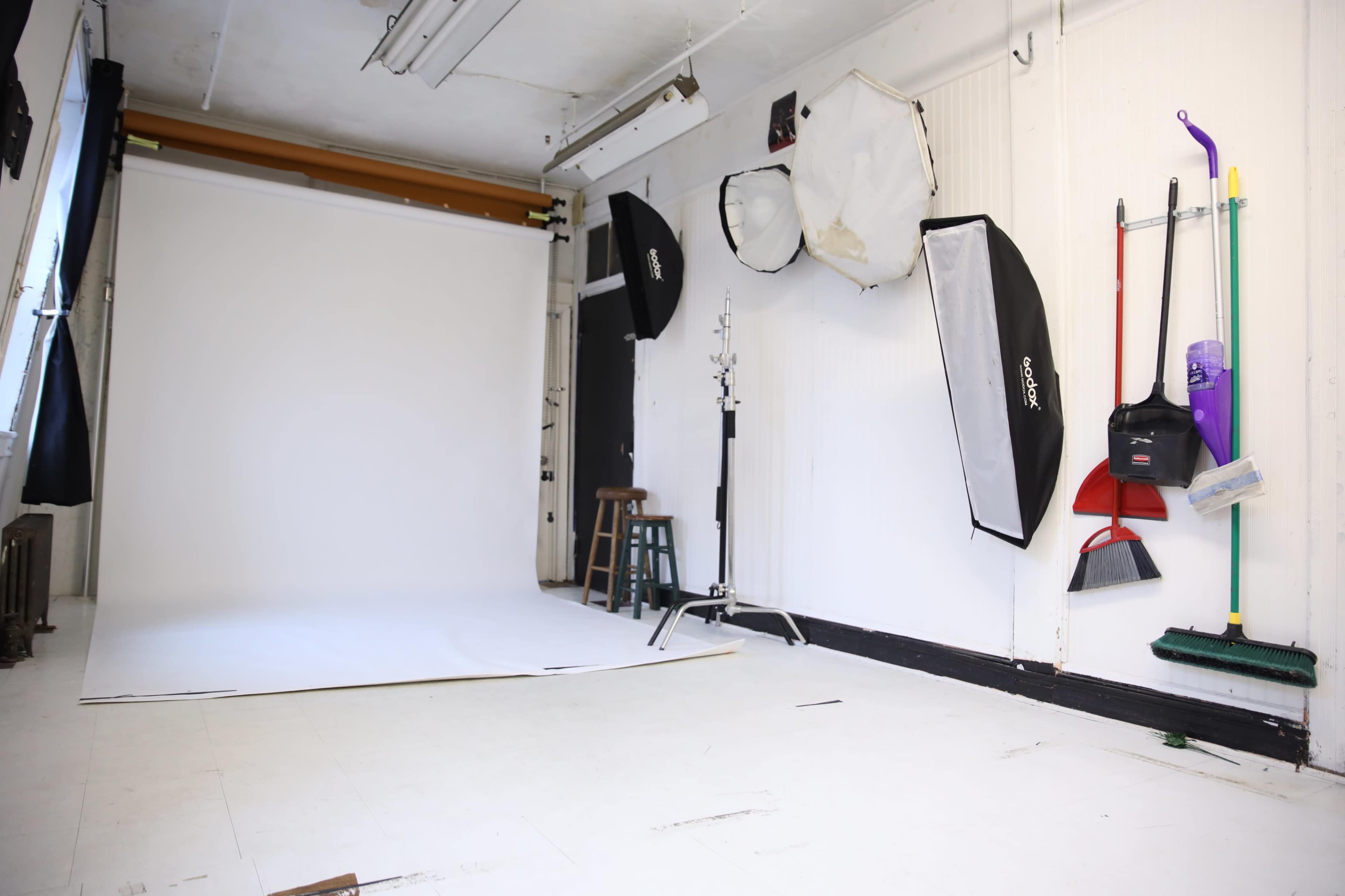 A photography studio with a white backdrop, various light modifiers, and cleaning tools hanging on the wall.