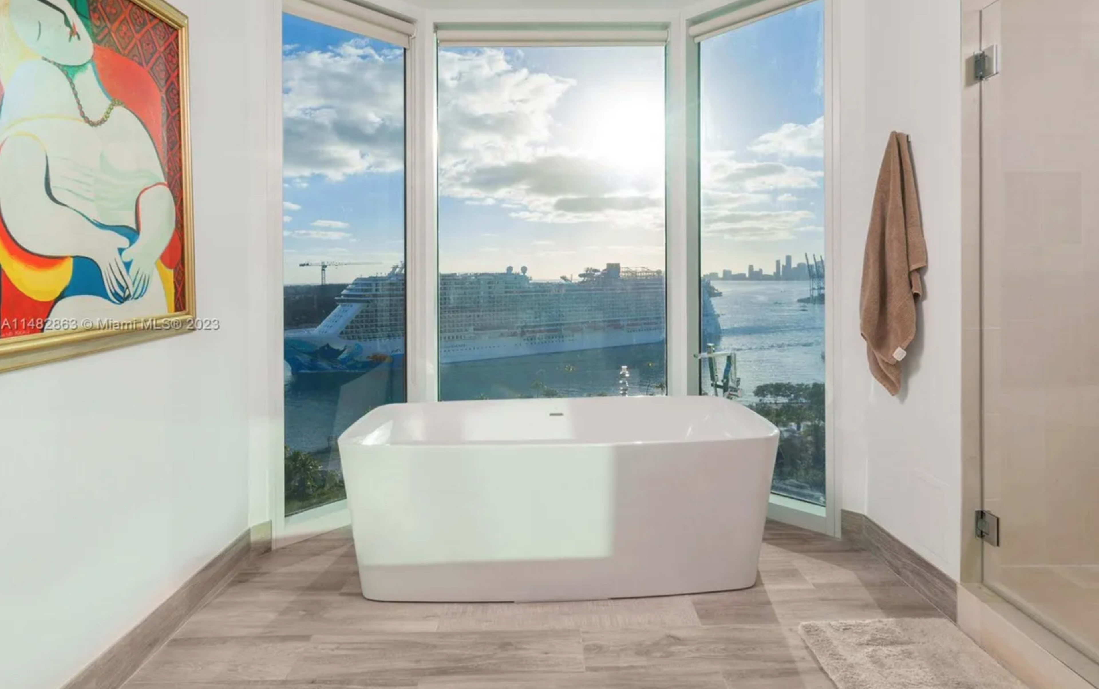 A modern freestanding bathtub is positioned by large windows overlooking a waterfront view with a cruise ship and city skyline in the background.