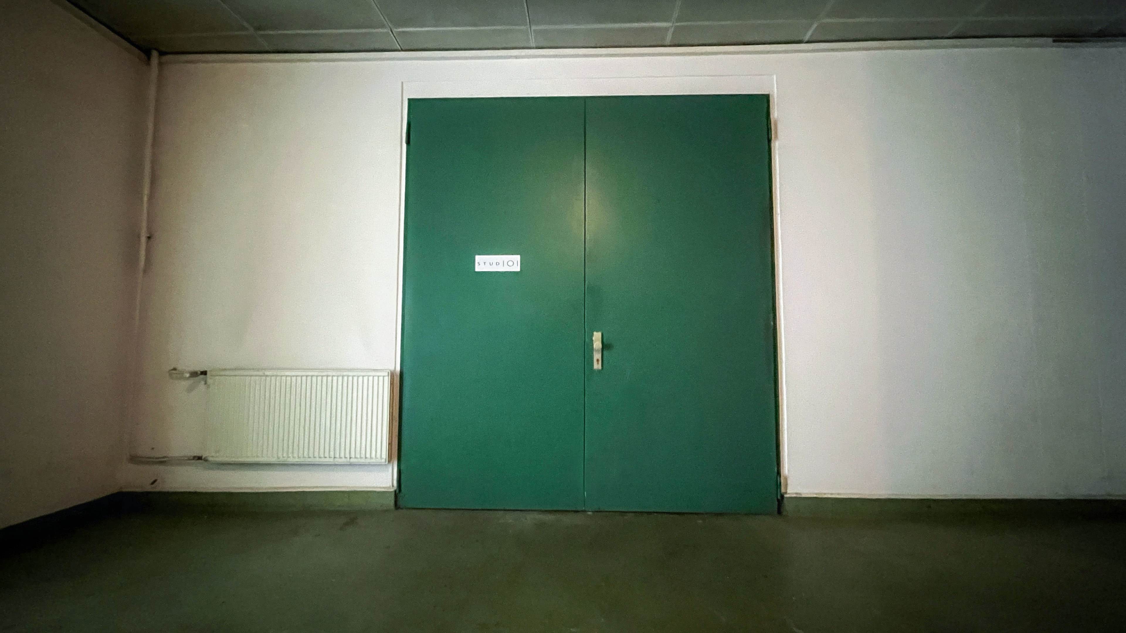 A pair of green double doors with a small sign stand against a plain wall, beside a white radiator.