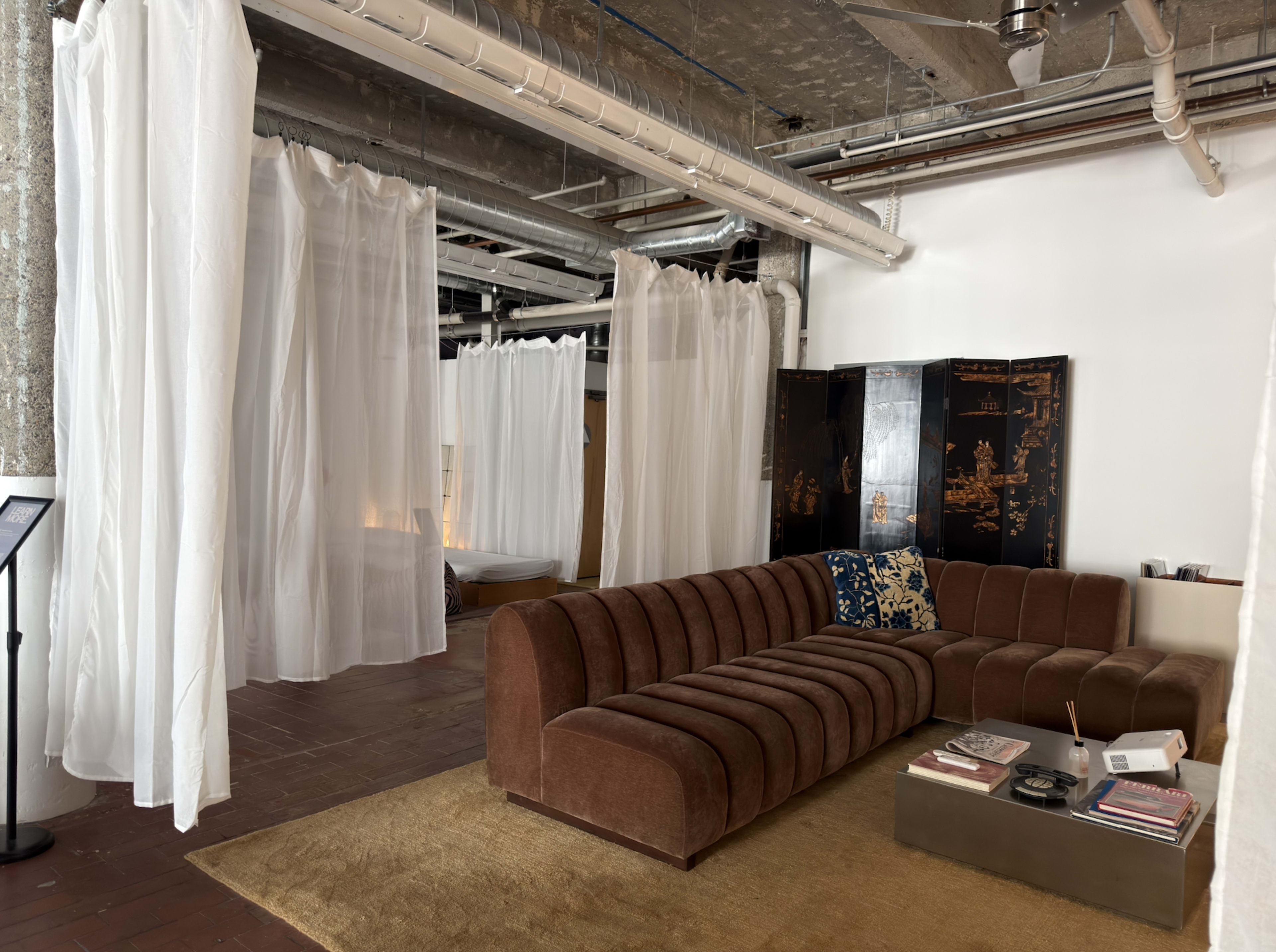 The image shows a modern interior space featuring a brown sectional sofa, light-colored curtains, and exposed industrial elements.