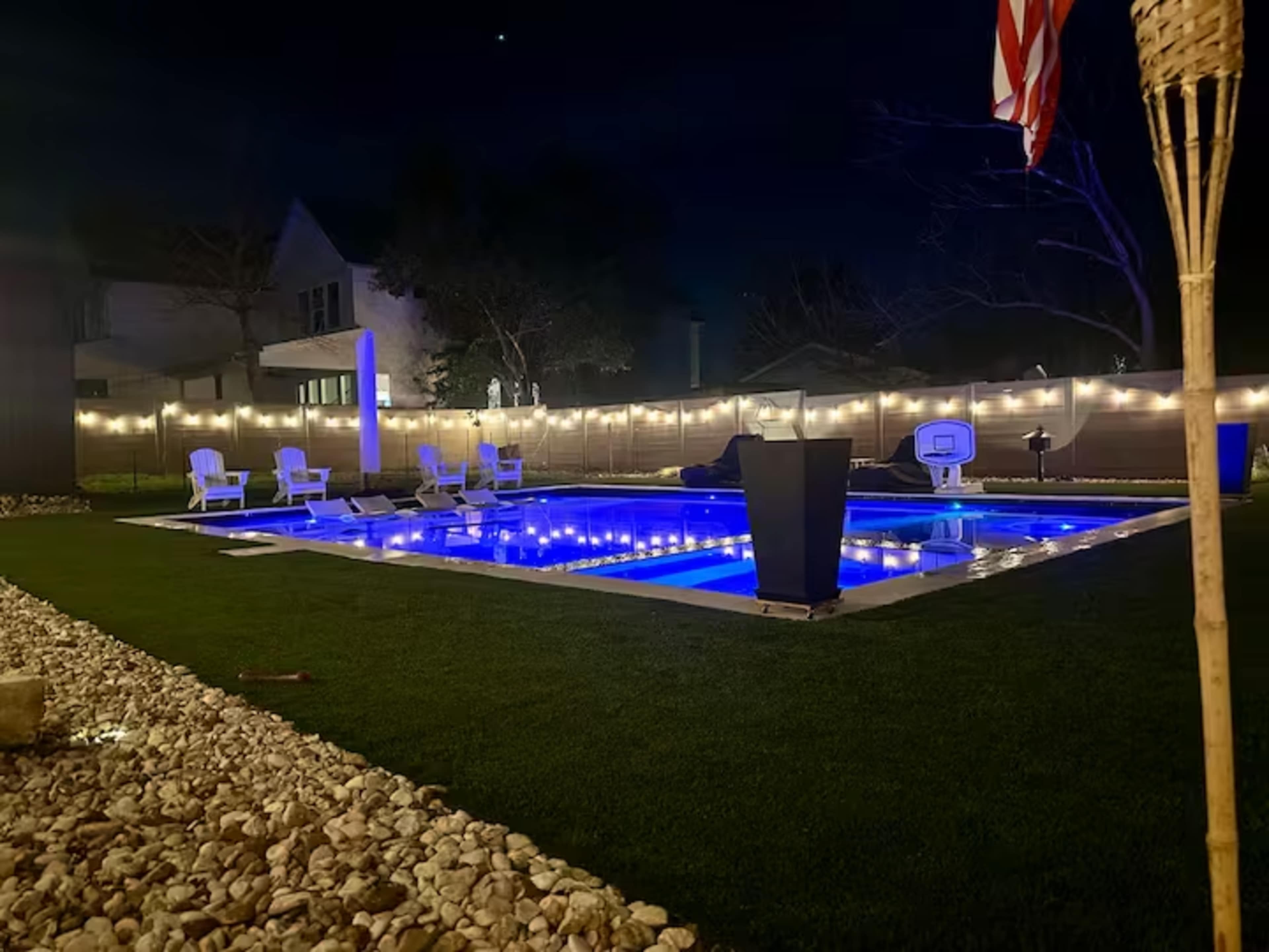 The image shows a well-lit backyard pool area at night, featuring blue pool lights, white lounge chairs, and decorative string lights.