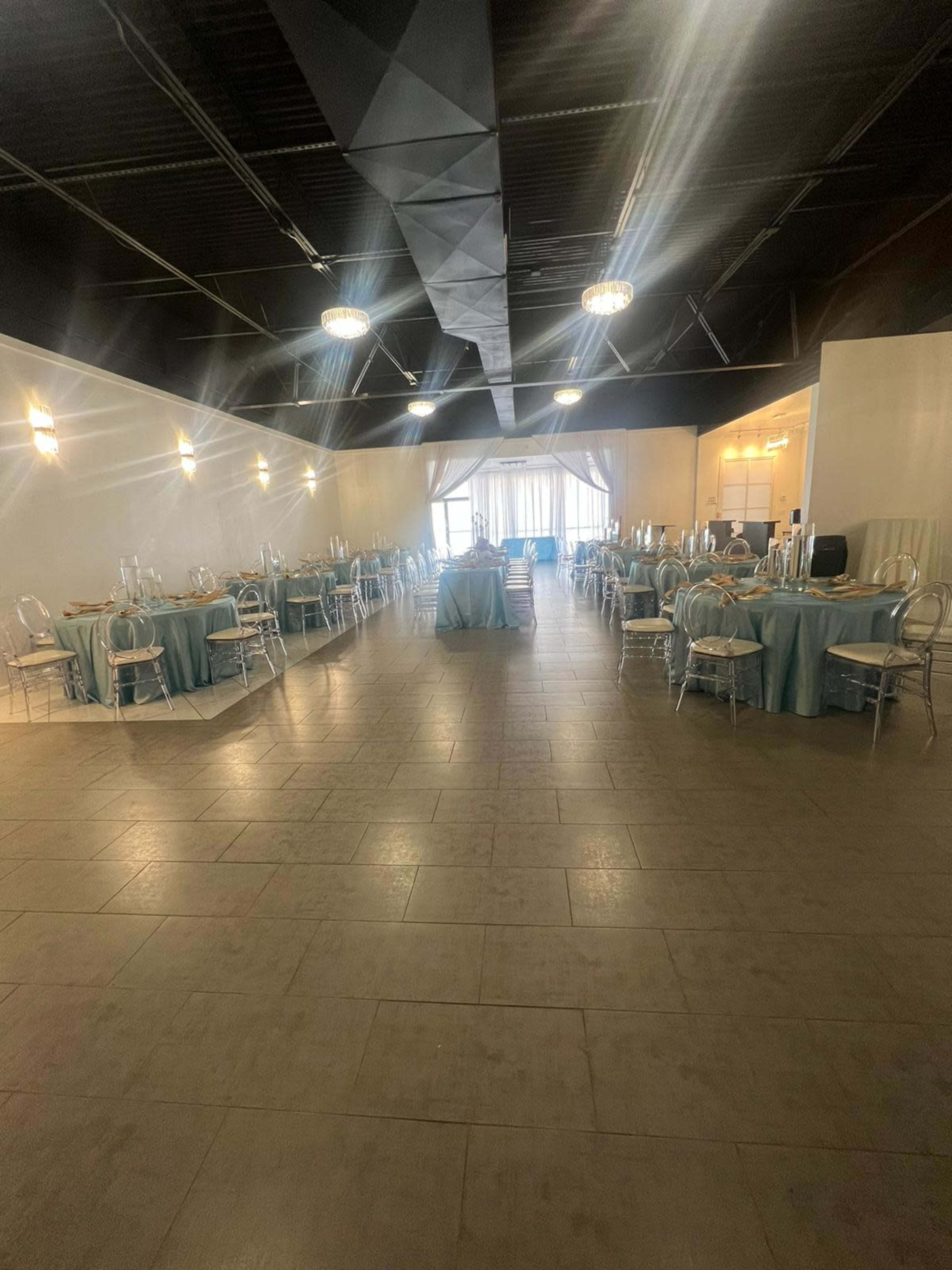 A spacious event room is set with circular tables covered in light blue tablecloths, arranged symmetrically around a central area that features a large window and a small serving counter.